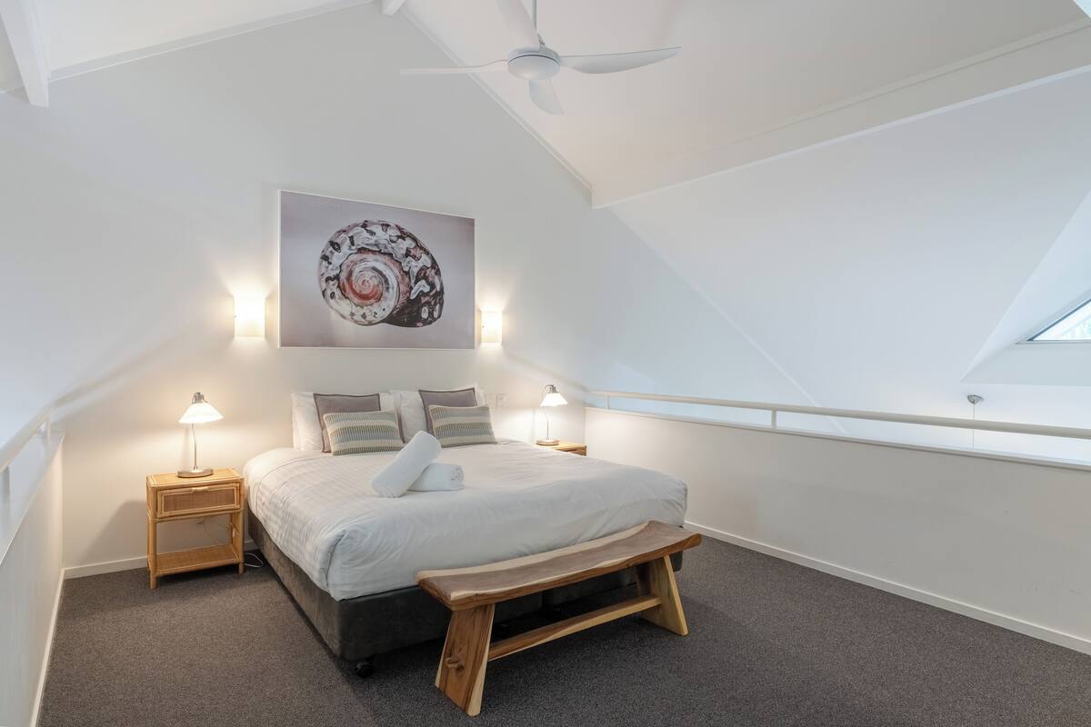 A bedroom with a large bed, bedside lamps, a wooden bench, a ceiling fan, and a skylight.