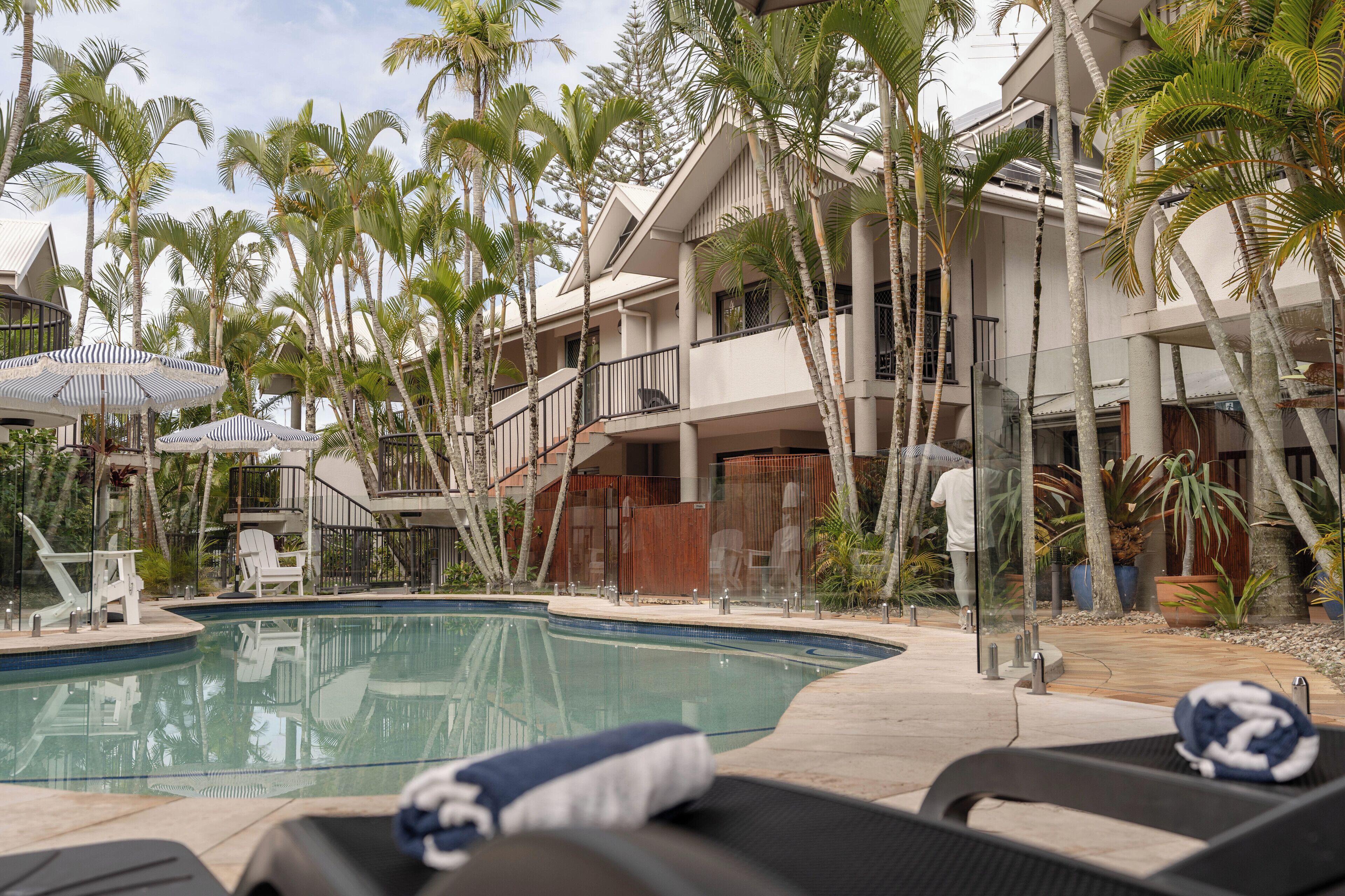 A pool area with lounge chairs, palm trees, and a building with balconies.