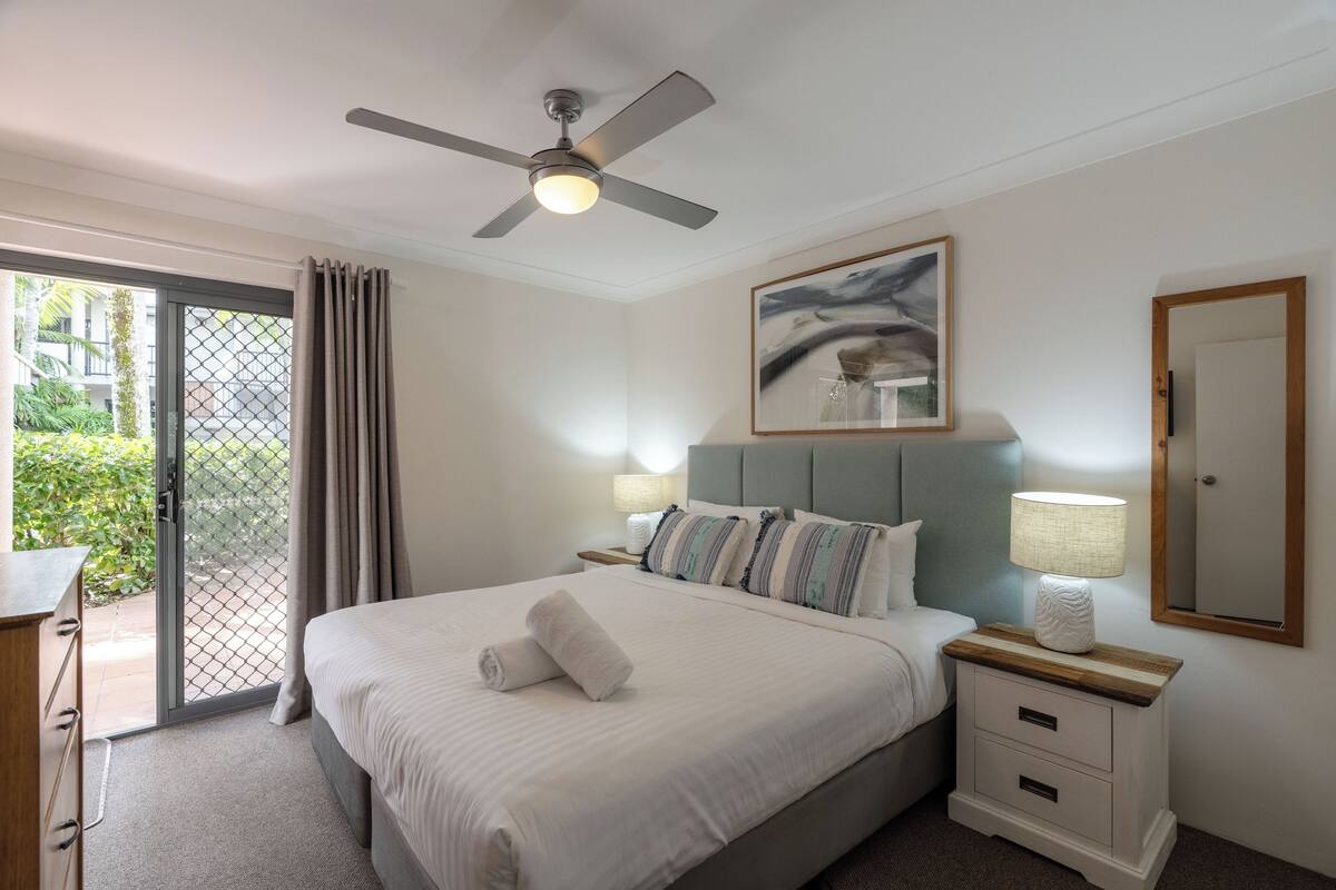 A hotel room with a bed, a ceiling fan, a nightstand with a lamp, a painting on the wall, and a sliding glass door leading to an outdoor area.