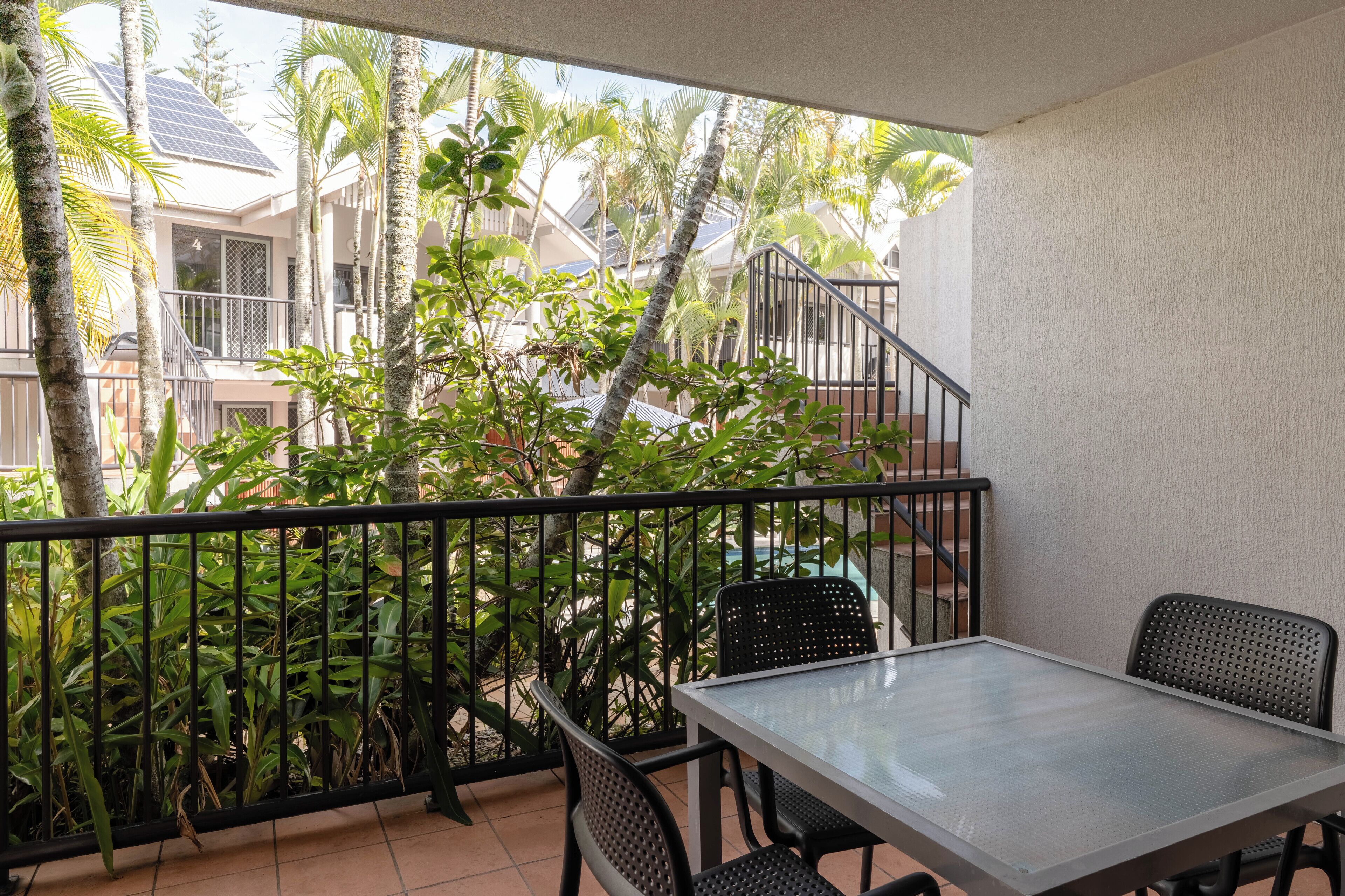 A balcony with a table and chairs, surrounded by lush greenery and a view of neighboring buildings.