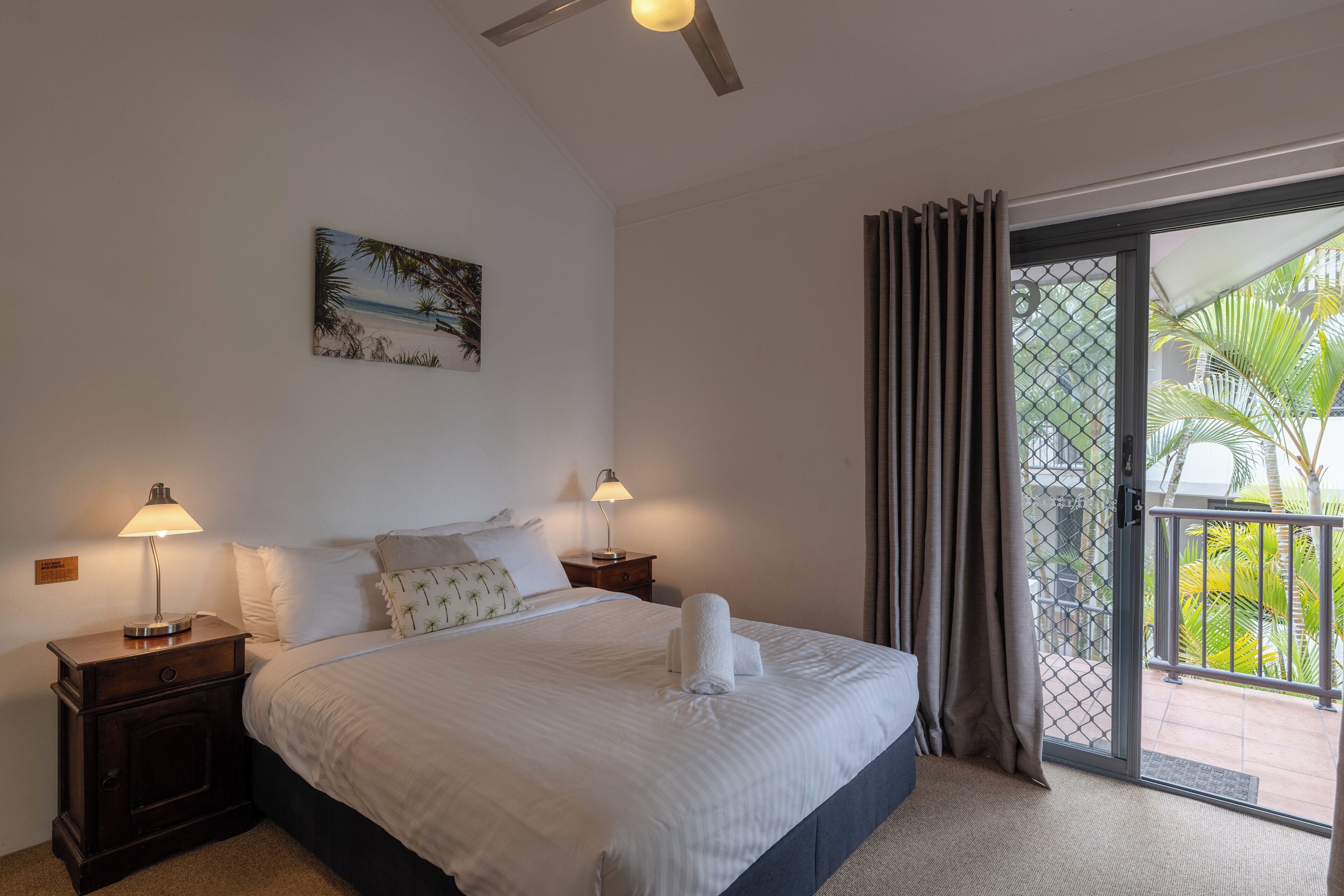 A neatly made bed with white linens and two bedside lamps, a painting of a beach scene, and a view of a balcony with palm trees.