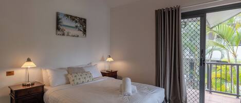 A neatly made bed with white linens and two bedside lamps, a painting of a beach scene, and a view of a balcony with palm trees.