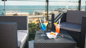 A balcony with wicker furniture, two drinks with fruit garnishes, and a view of the sea and buildings. - Aparthotel Sheila (Jesolo)