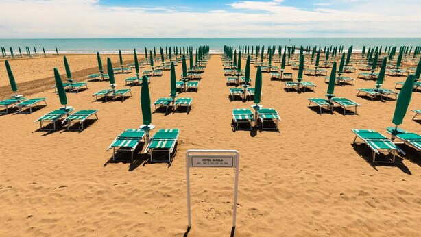 A beach with rows of green umbrellas and lounge chairs. A sign in the center reads "HOTEL SHEILA." - Aparthotel Sheila (Jesolo)