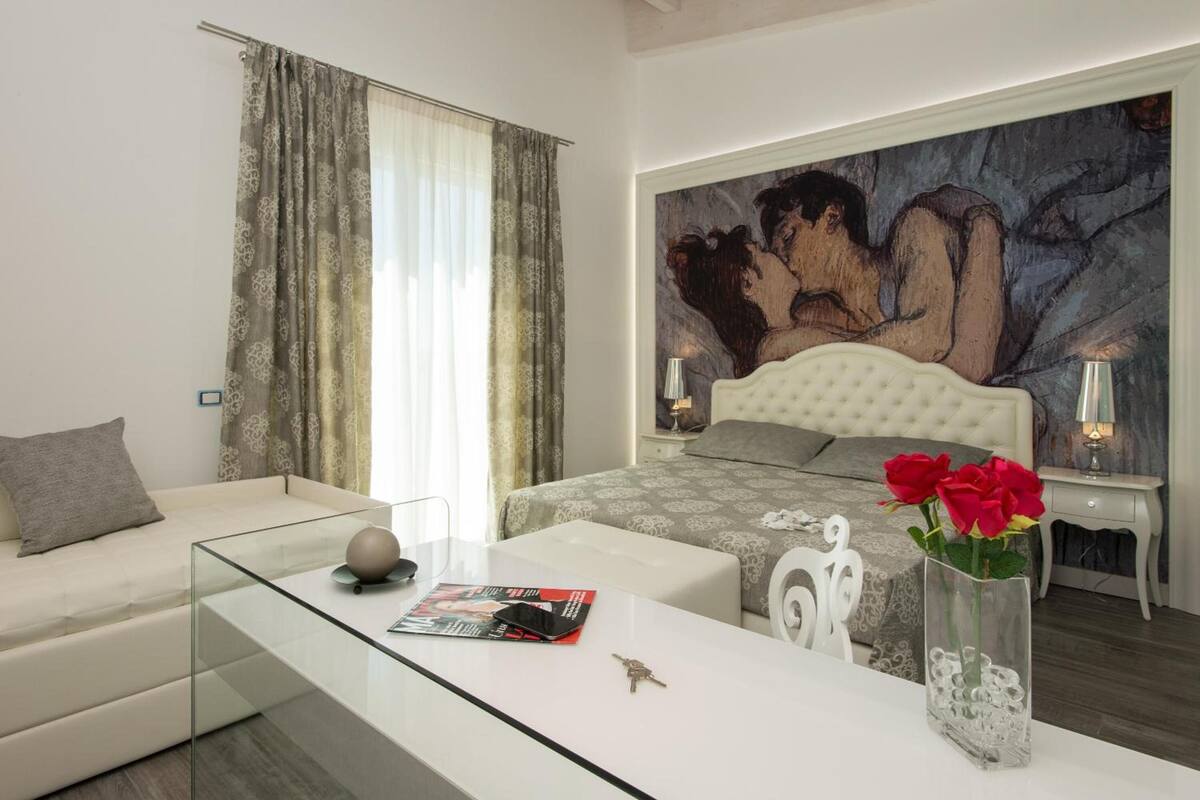A modern bedroom with a large bed, a glass coffee table, a white sofa, and a painting on the wall.