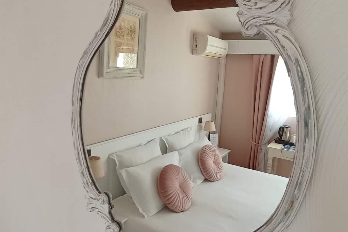 Ein Schlafzimmer mit einem großen, verzierten Spiegel, einem Bett mit weißen und rosafarbenen Kissen, einem Nachttisch mit Lampe und einem Fenster mit Vorhängen.