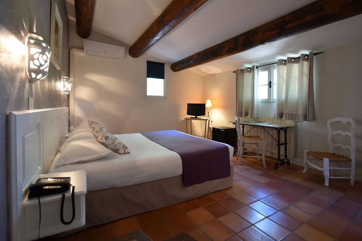 Ein Schlafzimmer mit Bett, Nachttisch, Lampe, Stuhl und Schreibtisch mit Computer.