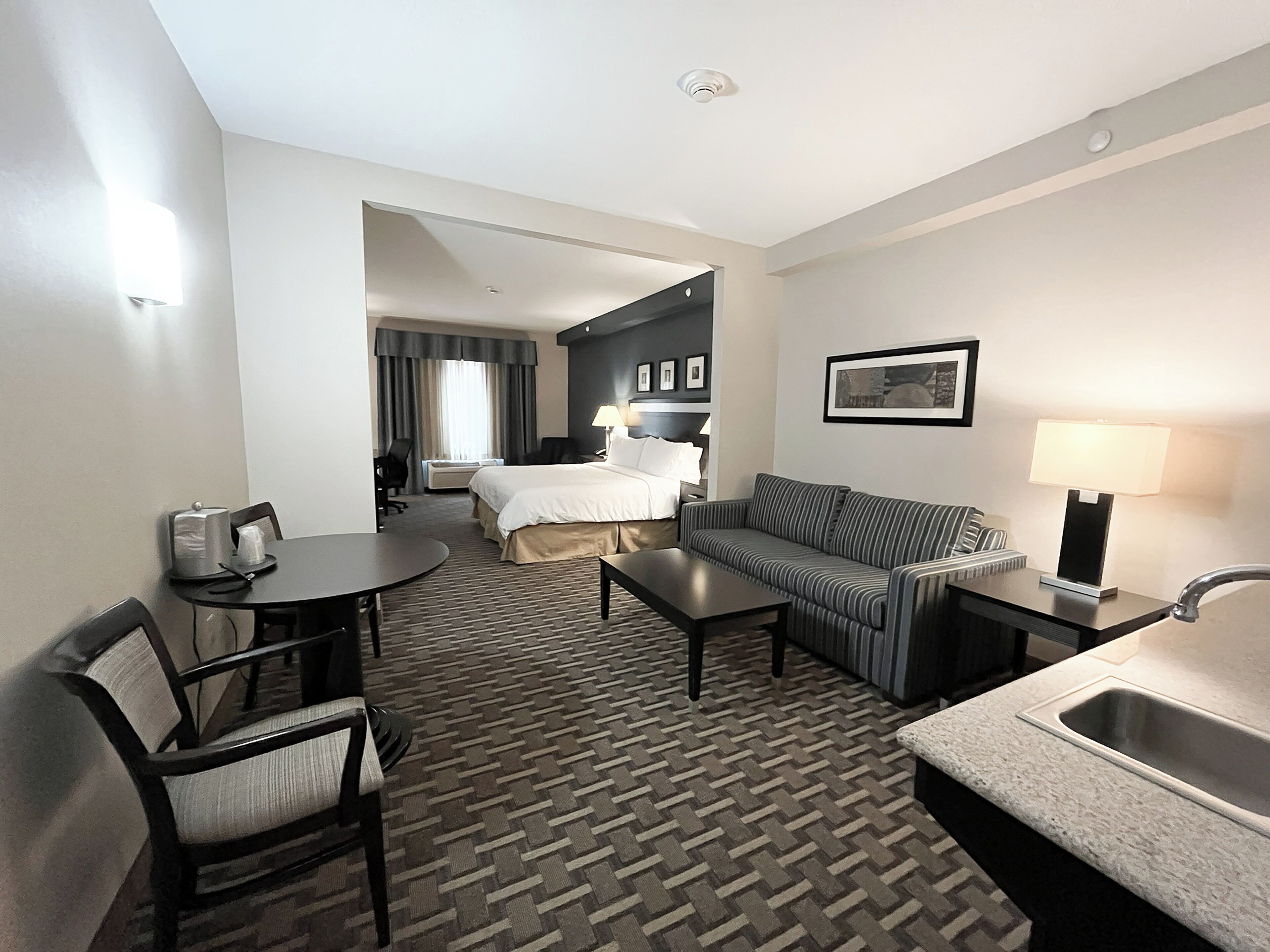 A hotel room with a bed, a sofa, a desk, a chair, a small table, a lamp, and a sink.