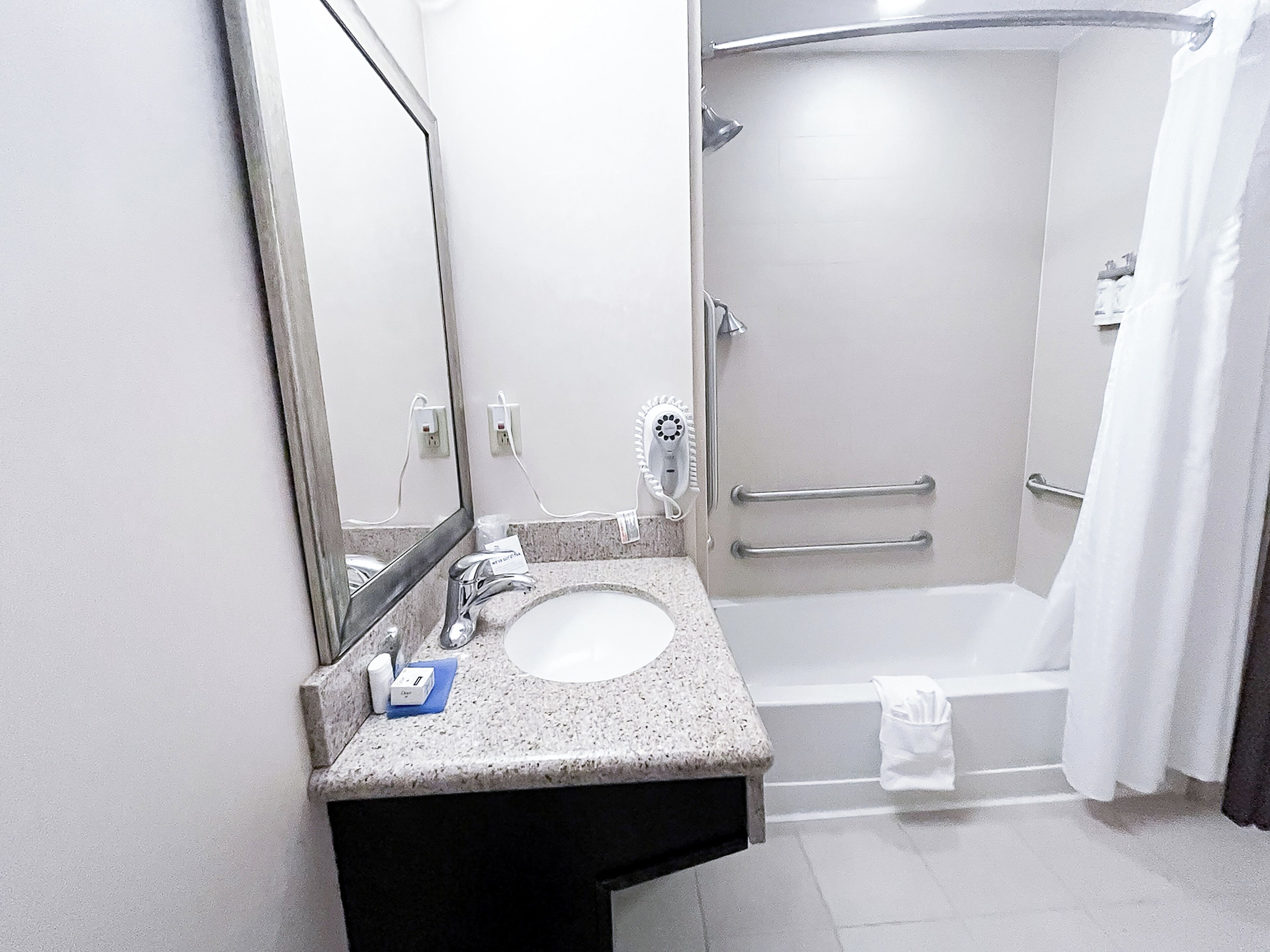 A modern bathroom with a granite countertop sink, a mirror, a handheld showerhead, and a walk-in shower with grab bars.