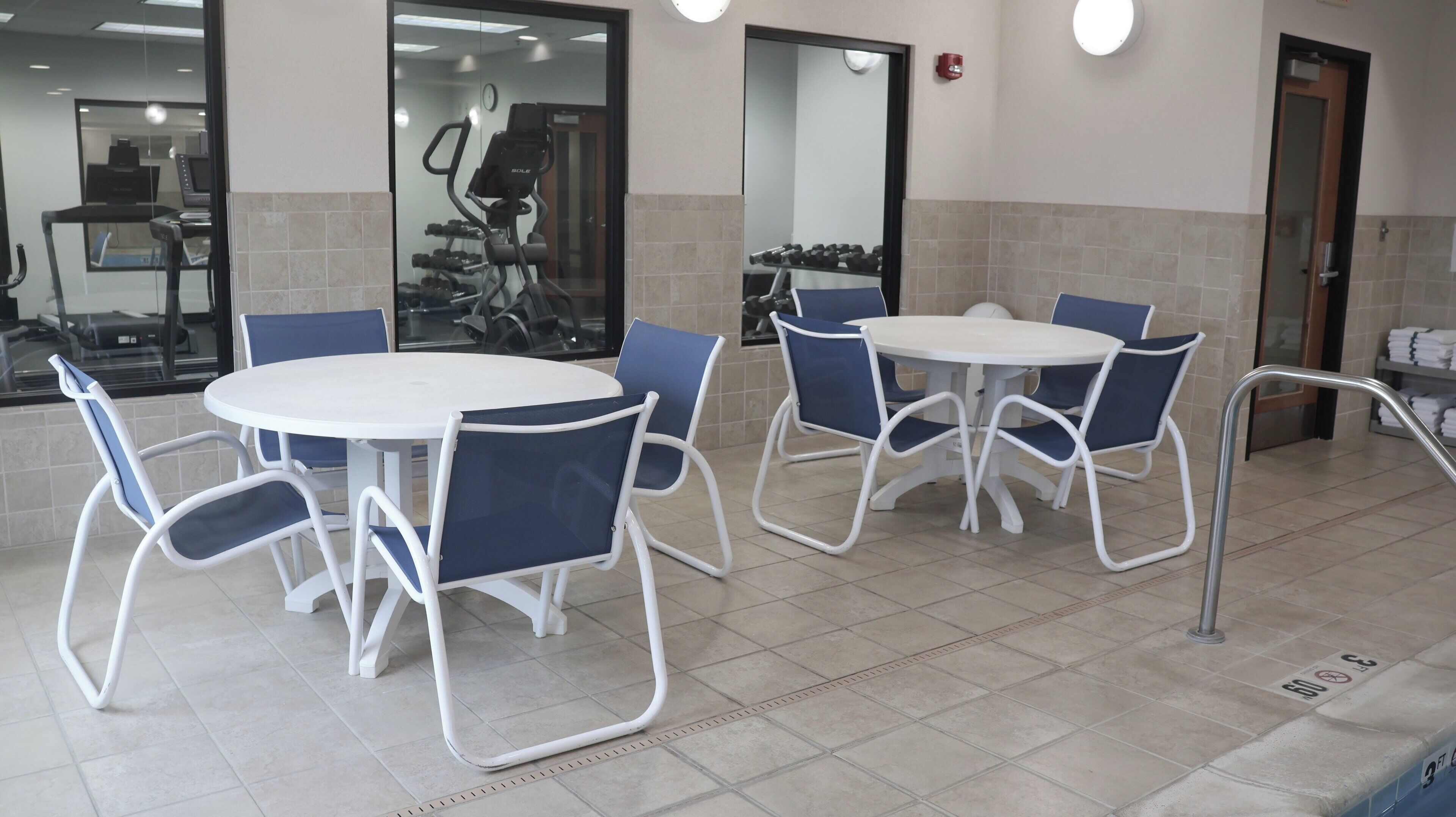 A dining area with round tables and blue chairs, a glass wall with exercise equipment, and a tiled floor.