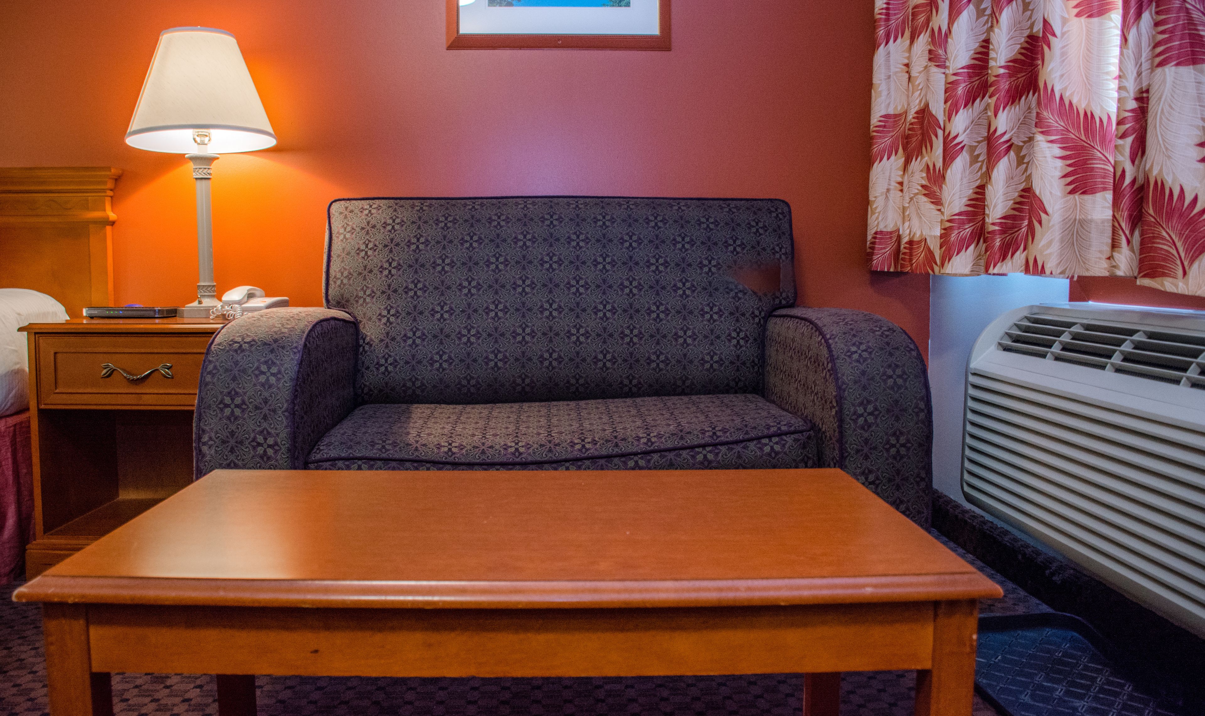 A hotel room with a single sofa, a wooden table, a lamp, a framed picture on the wall, and a window with curtains.