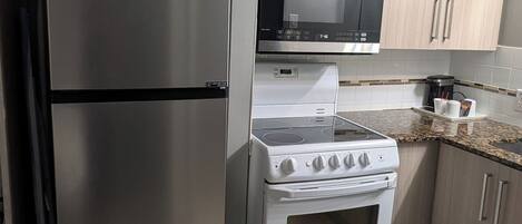 Una cocina compacta con un refrigerador de acero inoxidable, un microondas, un horno y alacenas de madera clara.