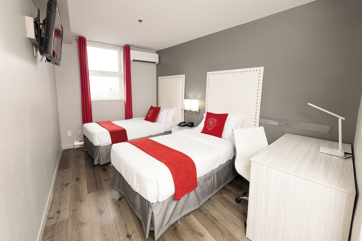 A hotel room with two beds, a desk, a TV, and a window with red curtains.