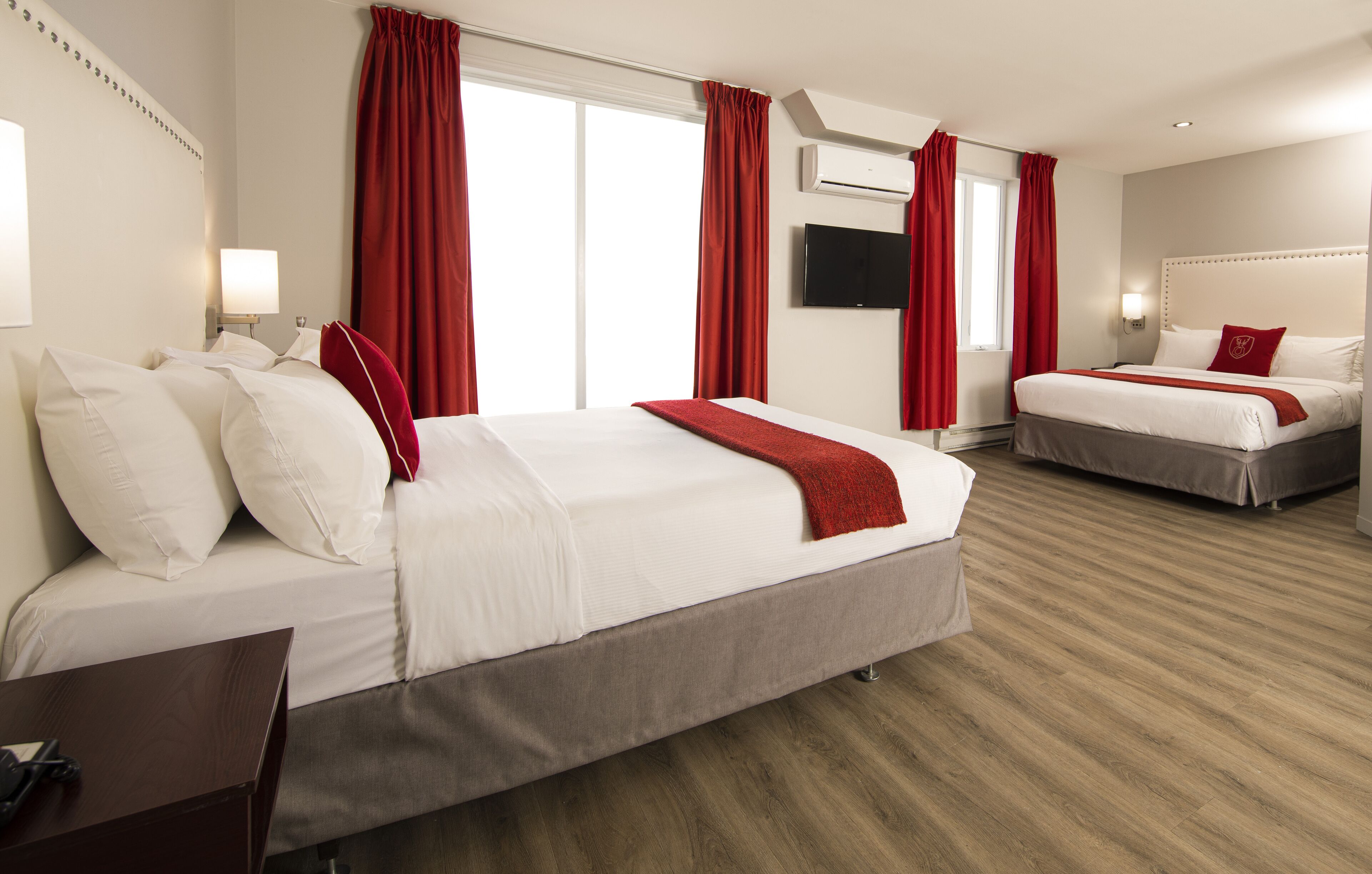 A hotel room with two beds, a desk, a TV, and a window with red curtains.