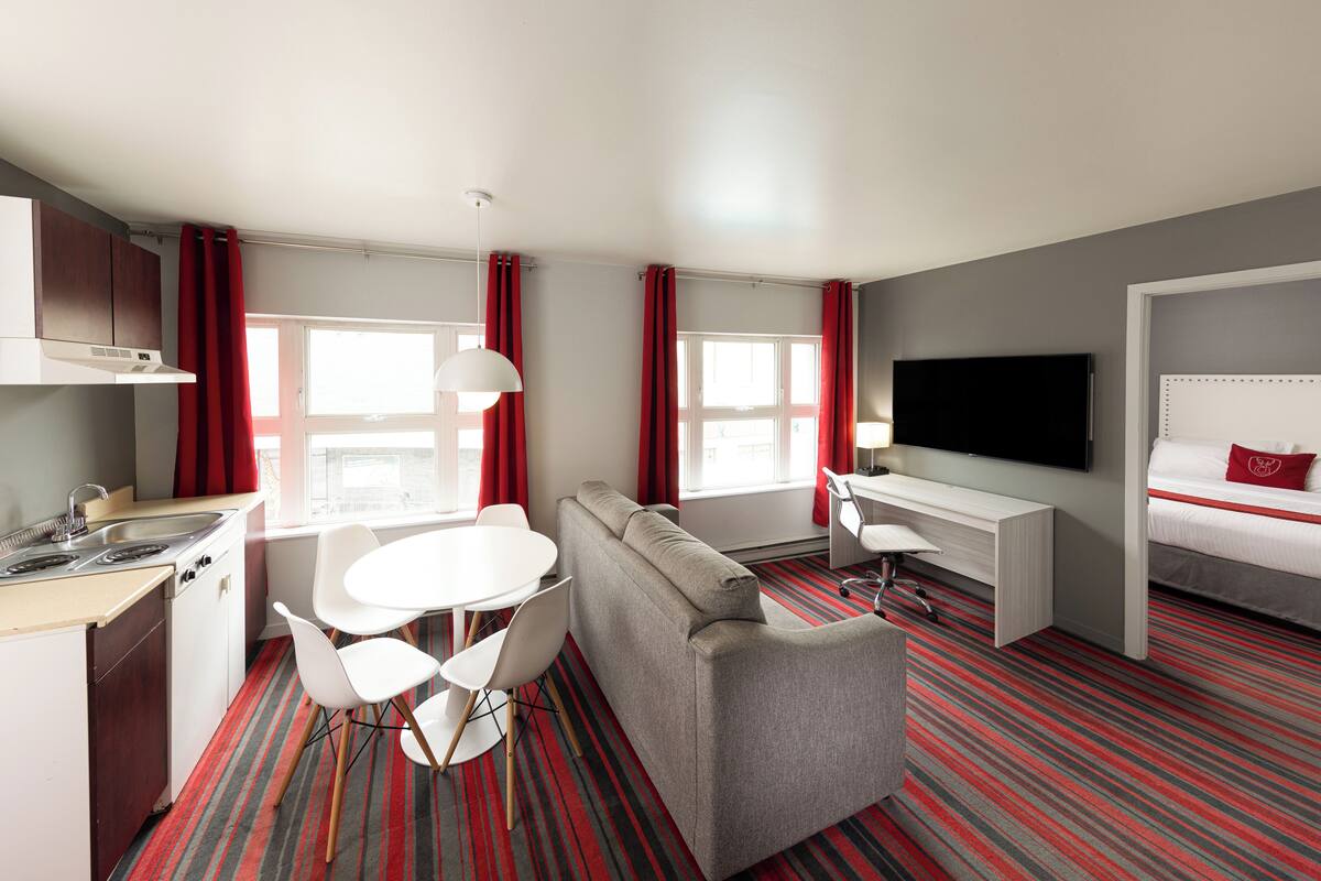 A compact hotel room with a kitchenette, a dining area with a table and chairs, a sofa, a desk with a chair, a TV mounted on the wall, and a bed with a red pillow.