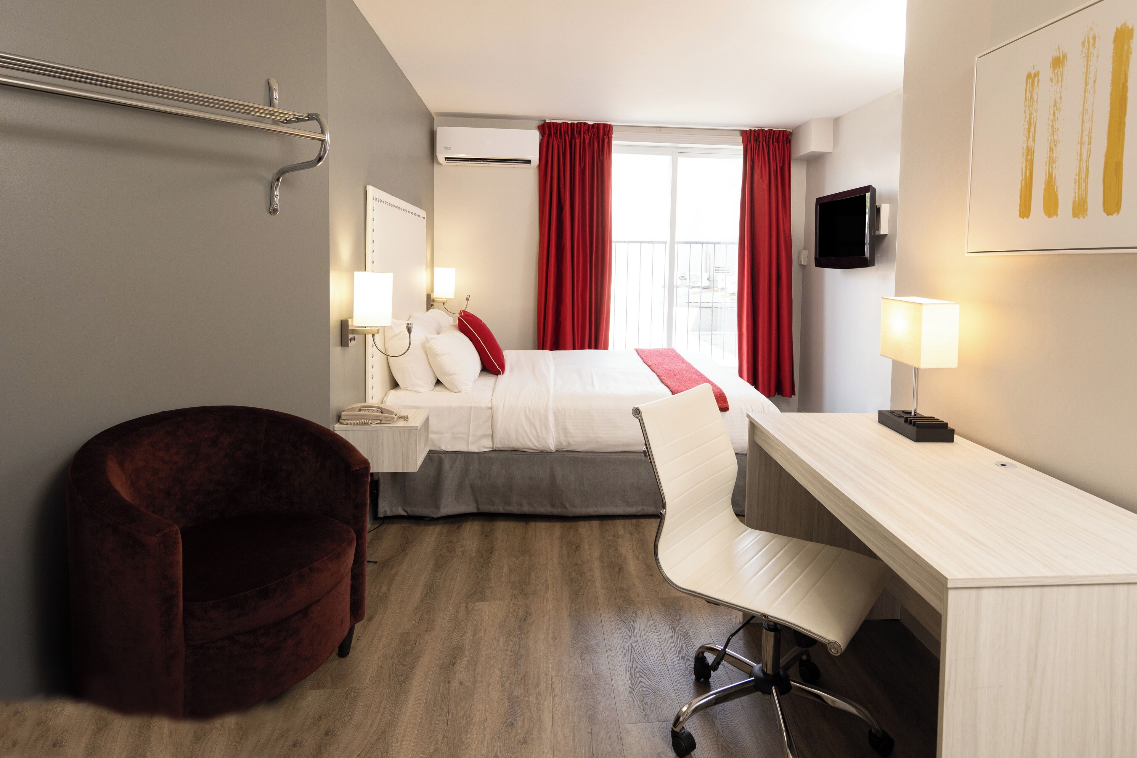A hotel room with a bed, a desk, a chair, a red velvet armchair, a TV, and a window with red curtains.