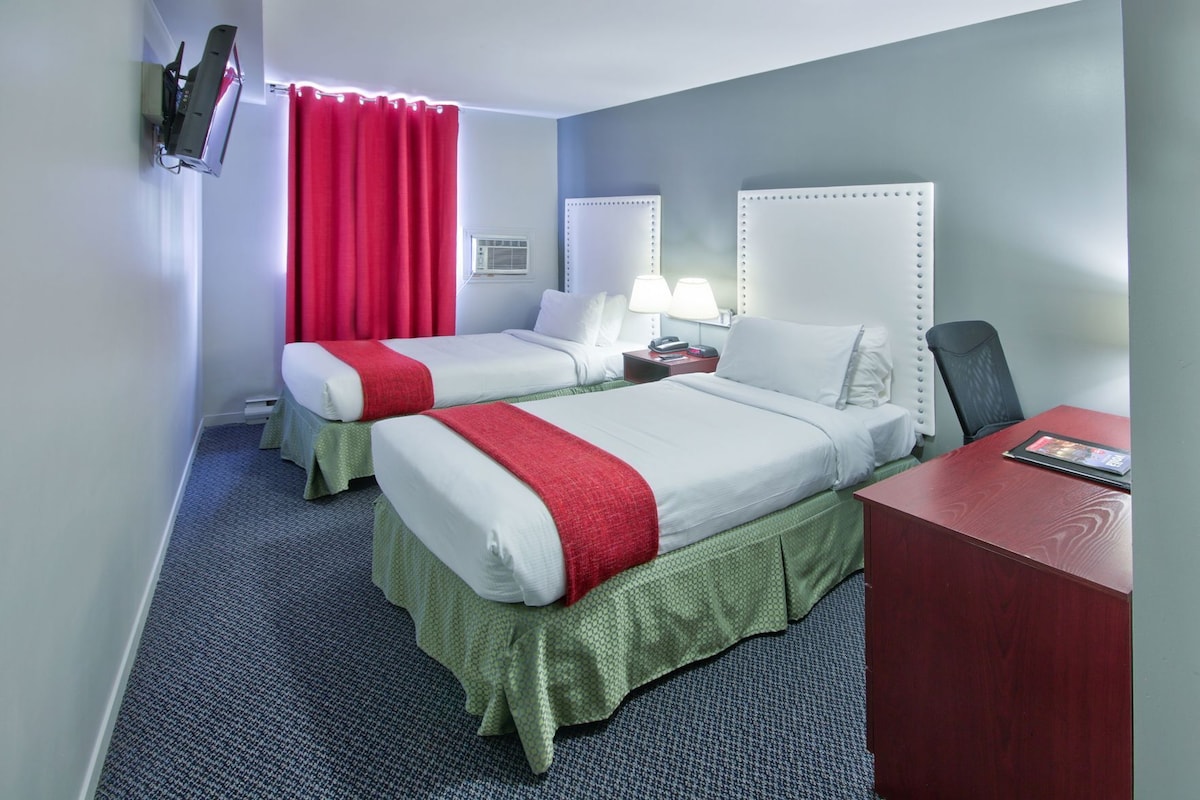 A hotel room with two beds, a desk, a chair, a TV mounted on the wall, and a window with curtains.