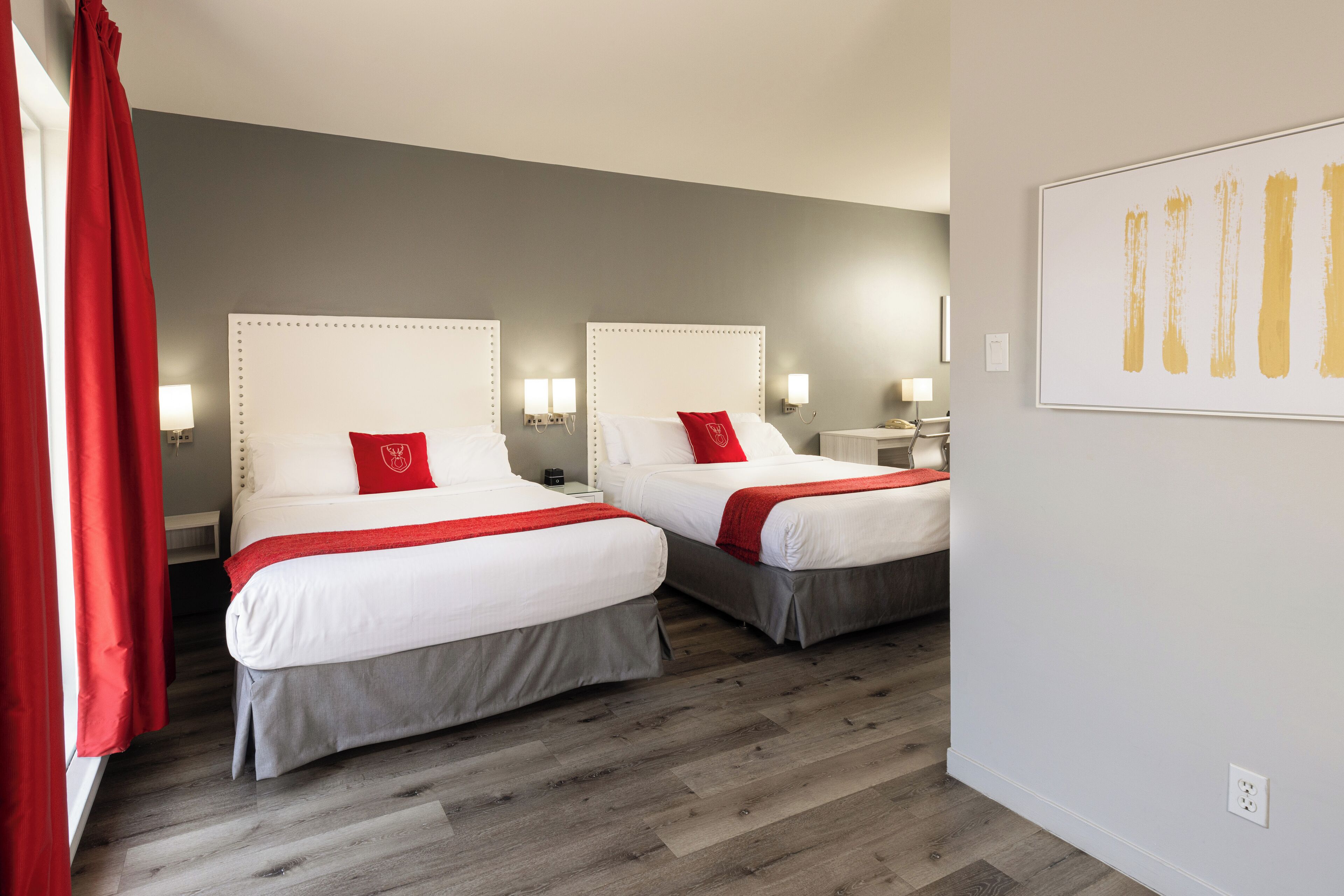Une chambre d’hôtel avec deux lits, chacun agrémenté d’un coussin rouge, d’une literie blanche et d’une couvre-lit grise. On y trouve des lampes de chevet et une œuvre d’art encadrée au mur.