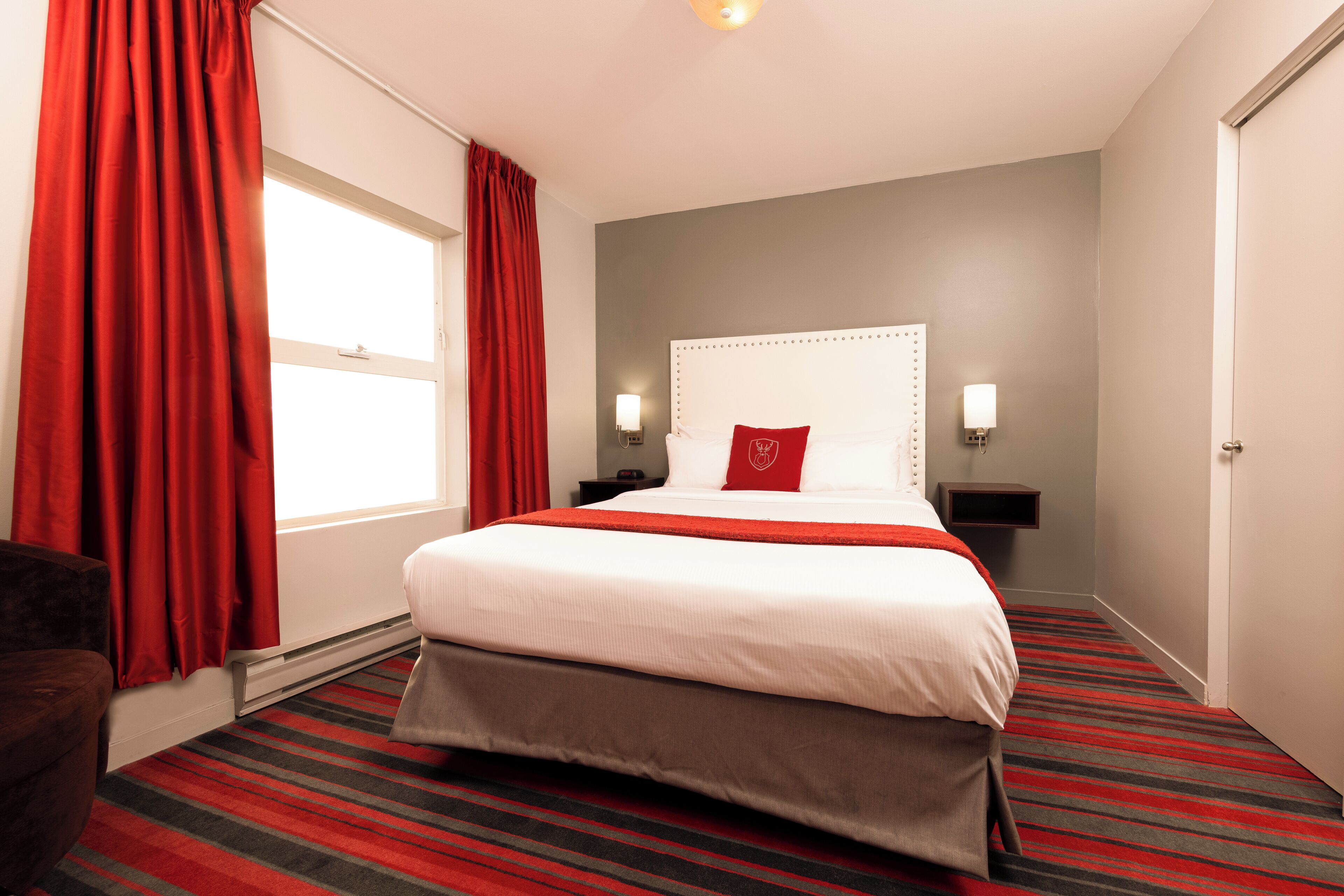 Una habitación de hotel con una cama grande, cortinas rojas, alfombra a rayas y un cabecero blanco.