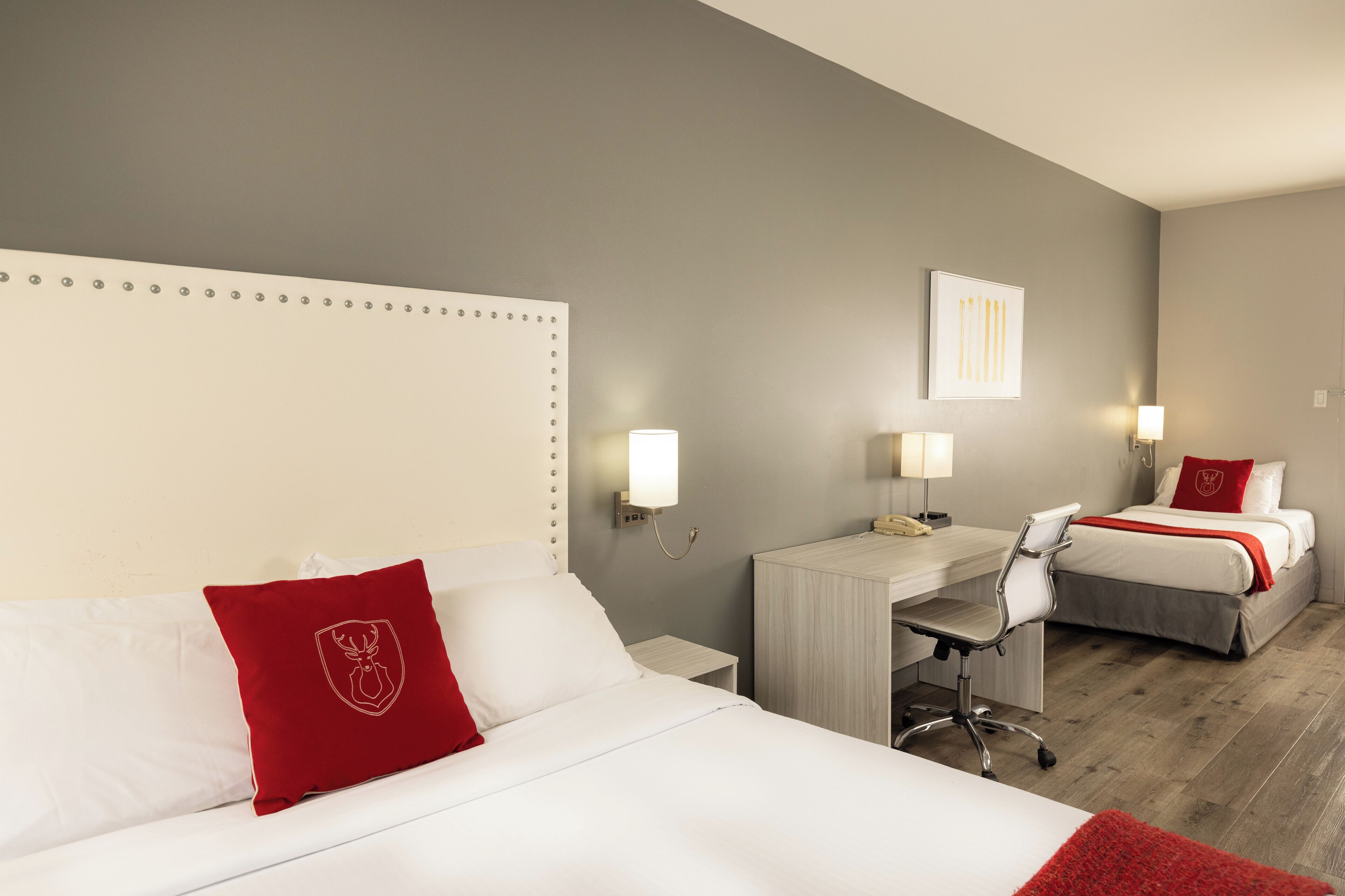 A hotel room with a large bed, a desk, and a smaller bed. There is a desk lamp, a chair, and a red pillow with a crest.
