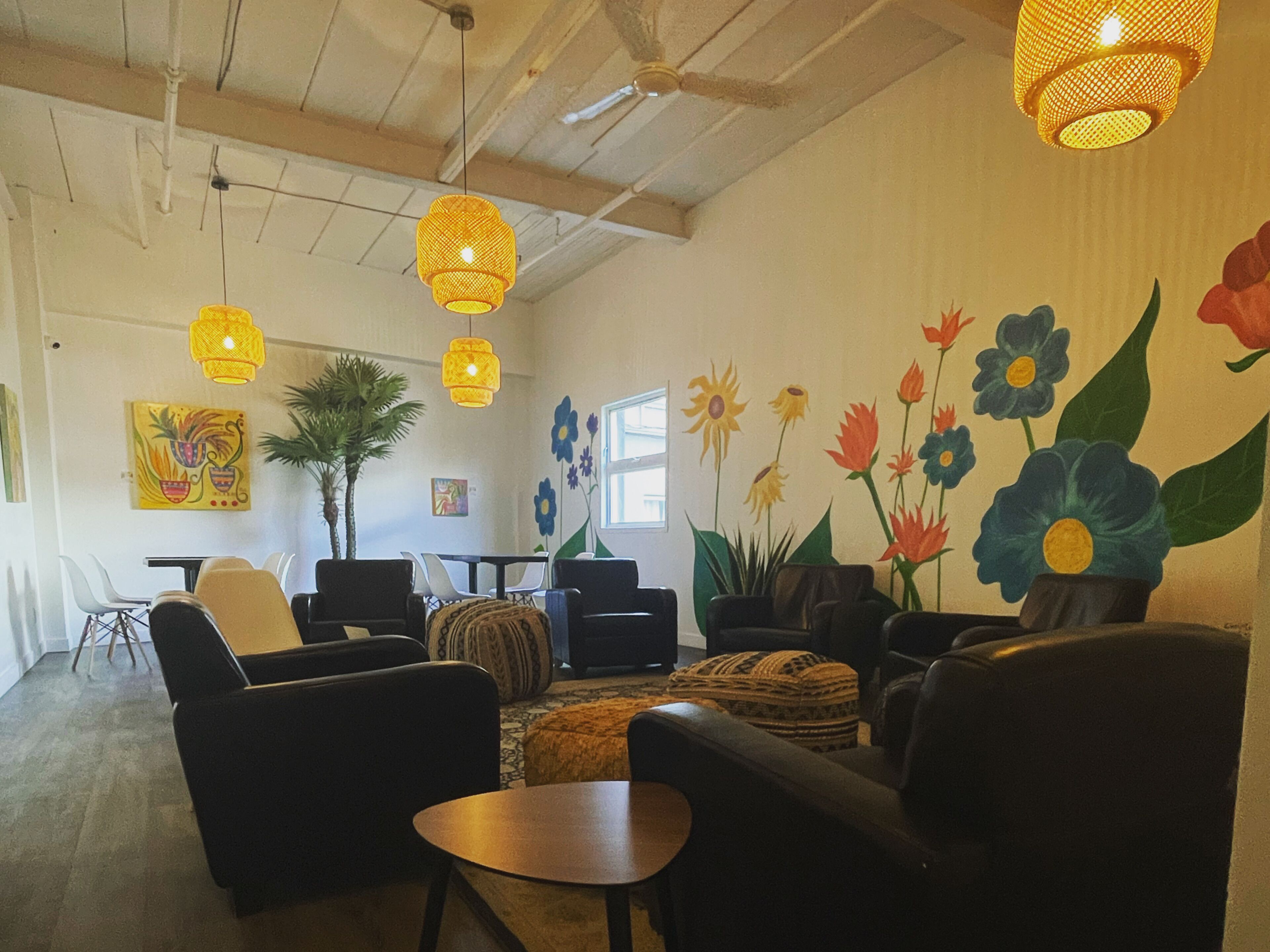 A spacious room with a high ceiling, featuring a large window, a dining area with chairs and a table, and a wall adorned with a floral mural.