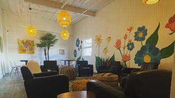 A spacious room with a high ceiling, featuring a large window, a dining area with chairs and a table, and a wall adorned with a floral mural.