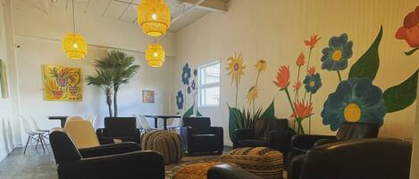 A spacious room with a high ceiling, featuring a large window, a dining area with chairs and a table, and a wall adorned with a floral mural.