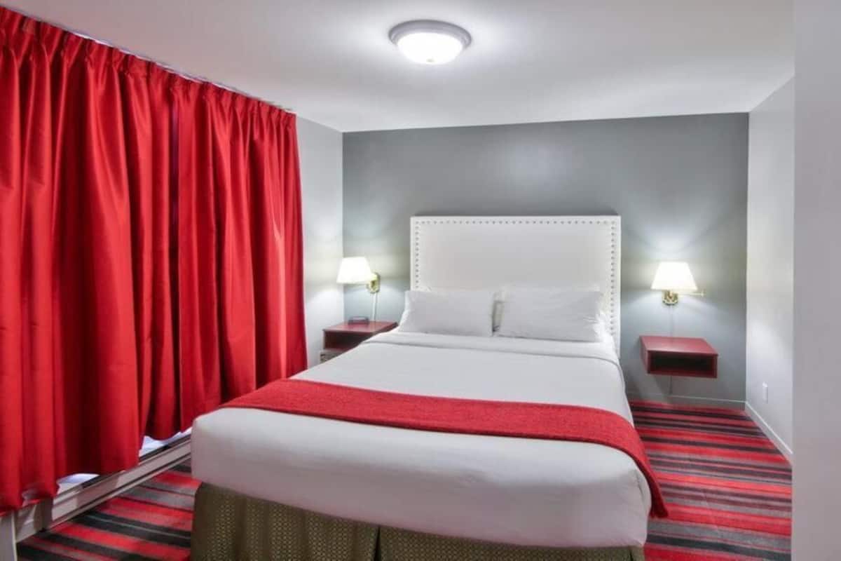 A hotel room with a bed, red curtains, two bedside lamps, and a red and black striped carpet.