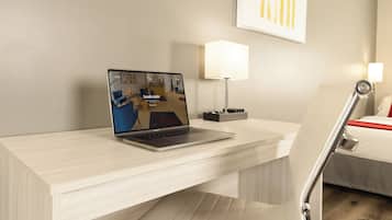 Quarto de hotel com um laptop em uma mesa, um abajur e uma cama com detalhes vermelhos.