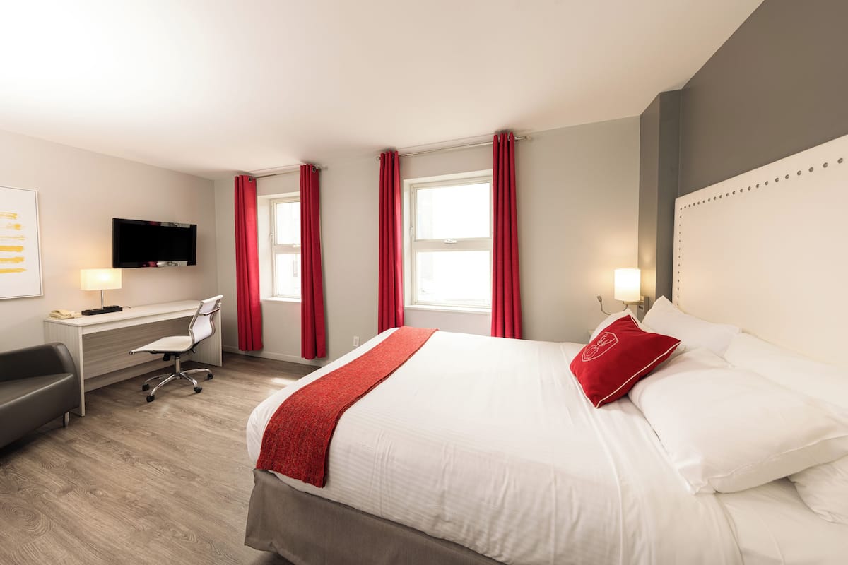 A hotel room with a bed, desk, chair, TV, and windows with red curtains.