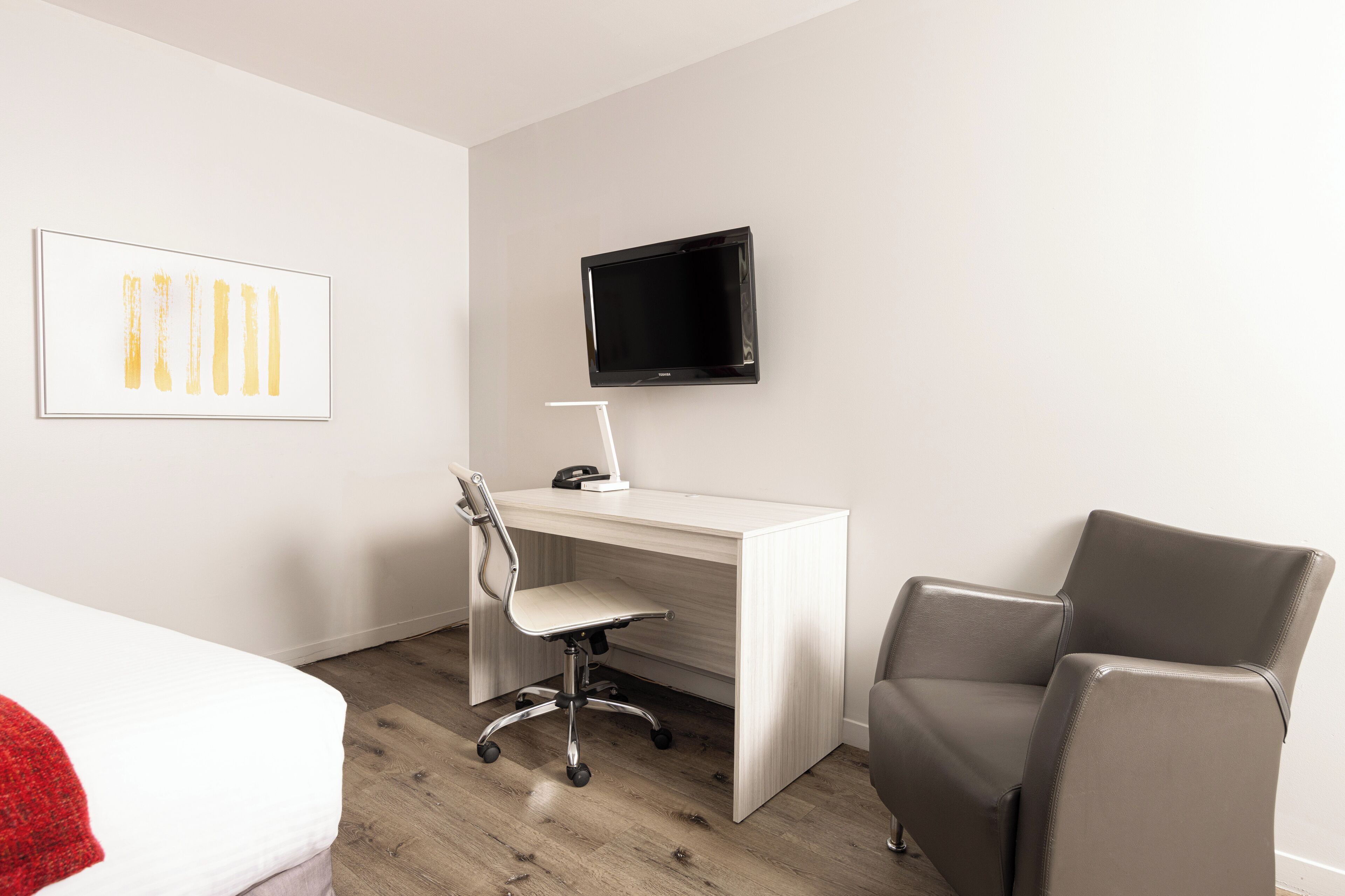 A hotel room with a bed, a desk with a chair, a TV mounted on the wall, and a framed picture on the wall.