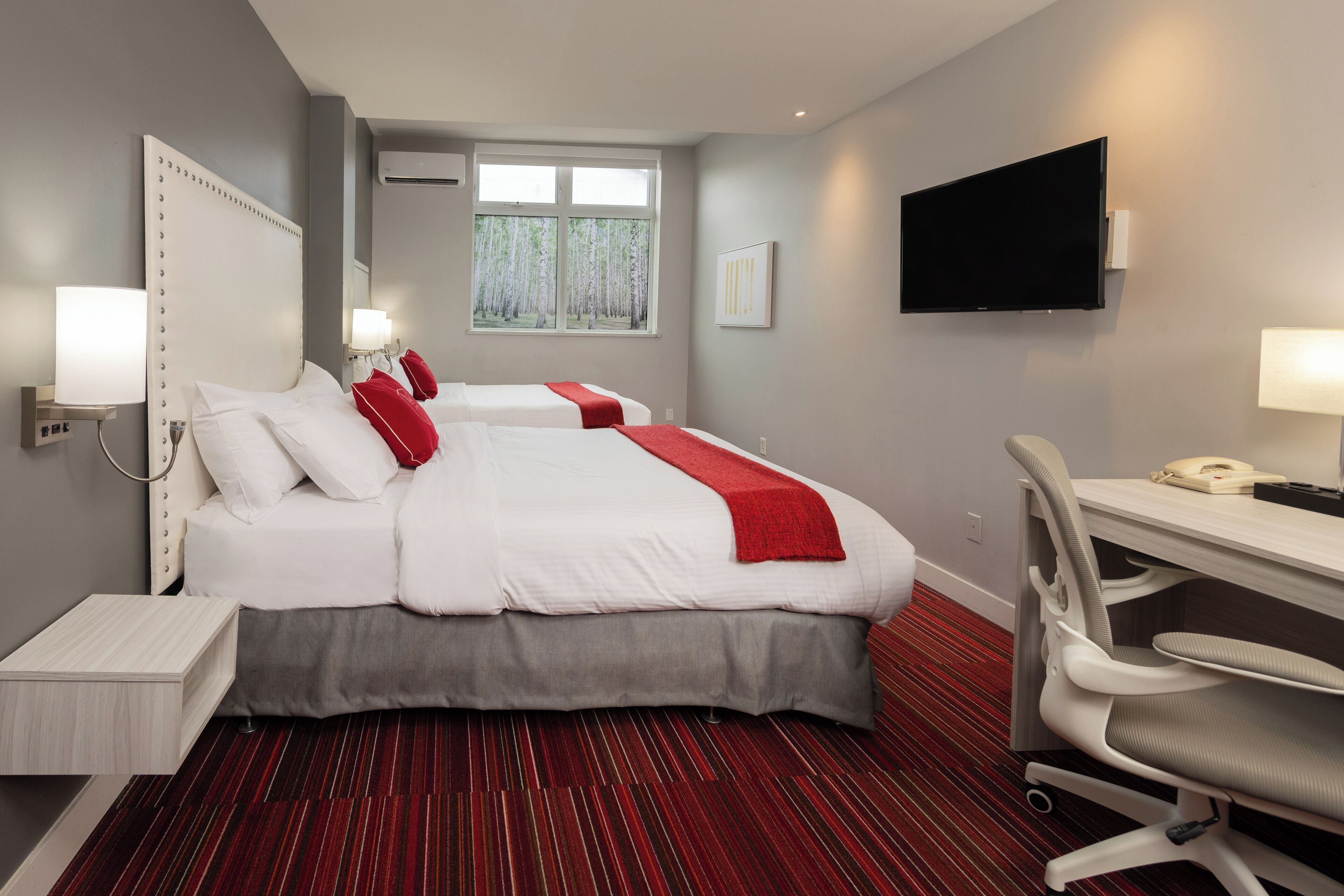 A hotel room with two beds, a desk, a chair, a TV, and a window with a view of trees.