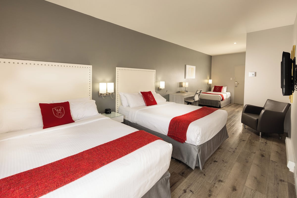 A hotel room with three beds, each with a red accent pillow, white bedding, and a red throw. There is a desk with a lamp, a chair, and a TV mounted on the wall.