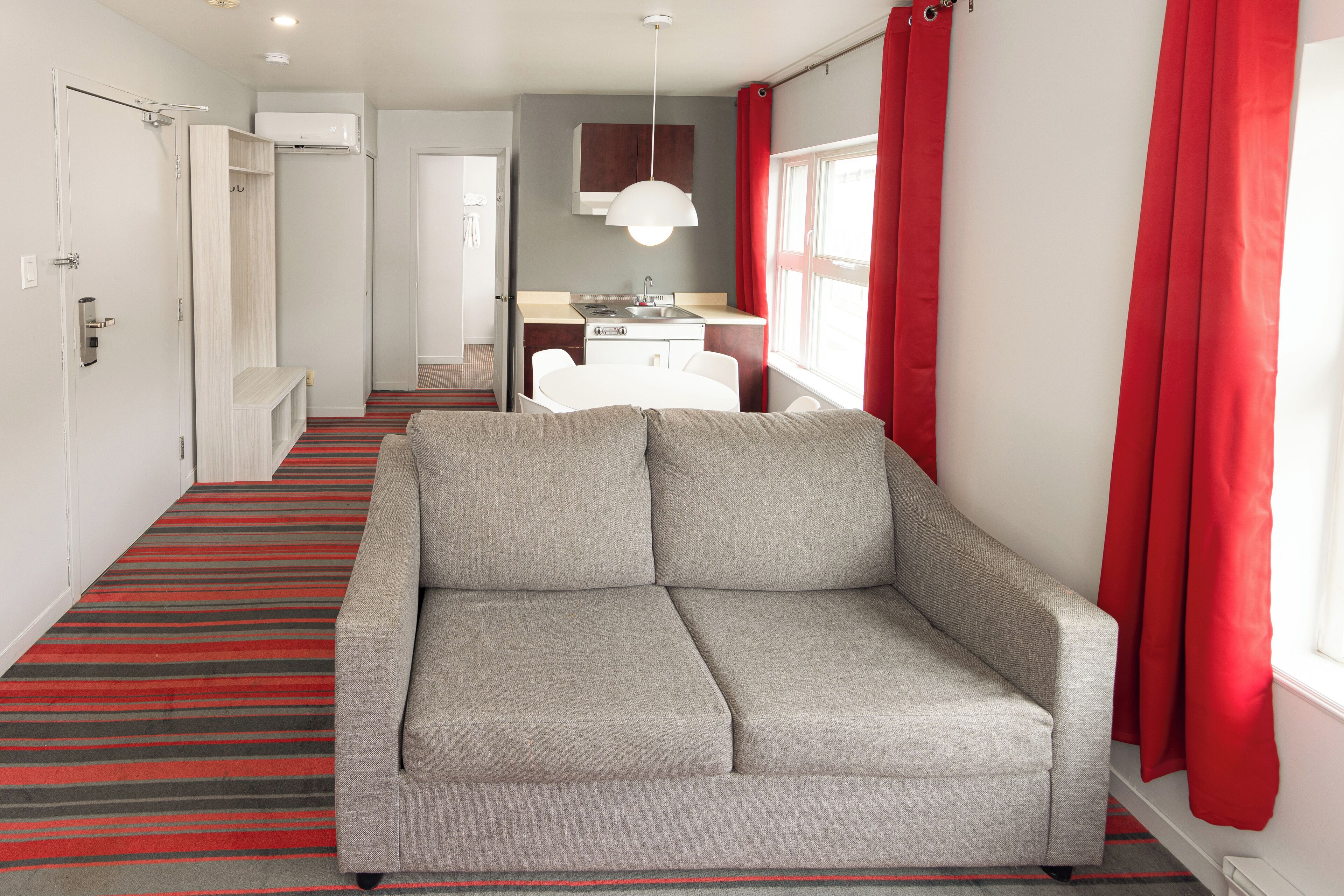 Una habitación de hotel moderna con sofá gris, alfombra a rayas rojas, una pequeña kitchenette y una ventana con cortinas rojas.