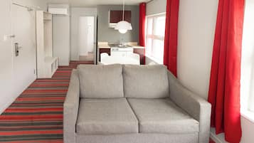 Una habitación de hotel moderna con sofá gris, alfombra a rayas rojas, una pequeña kitchenette y una ventana con cortinas rojas.