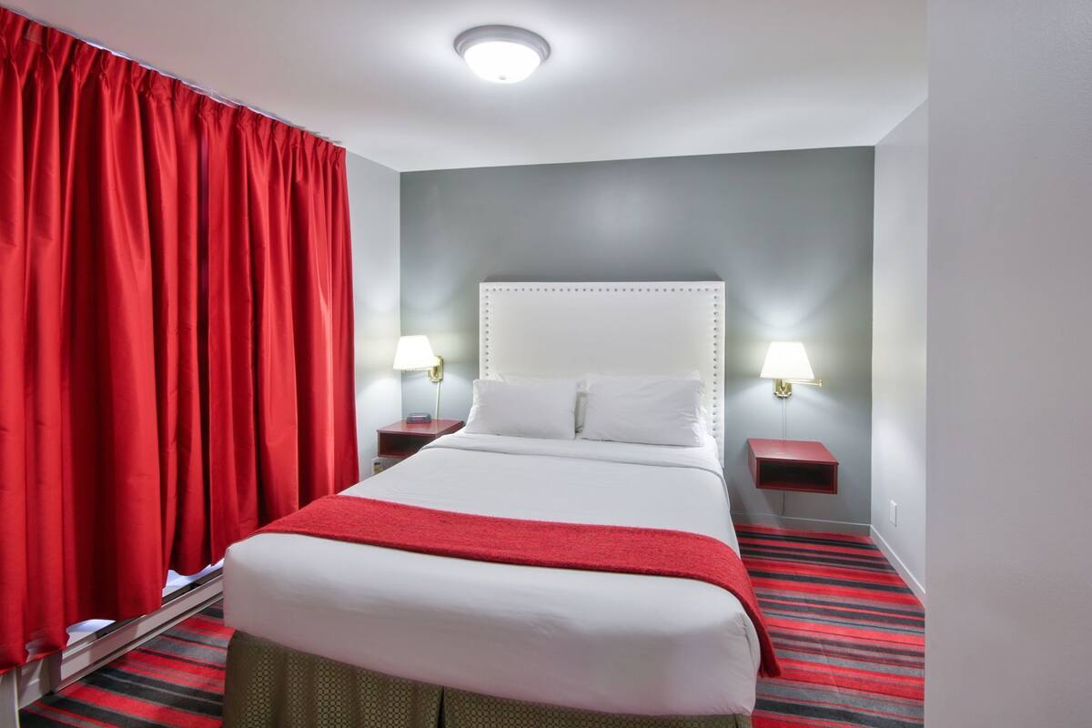 A hotel room with a bed, red curtains, two bedside lamps, and a red and black striped carpet.