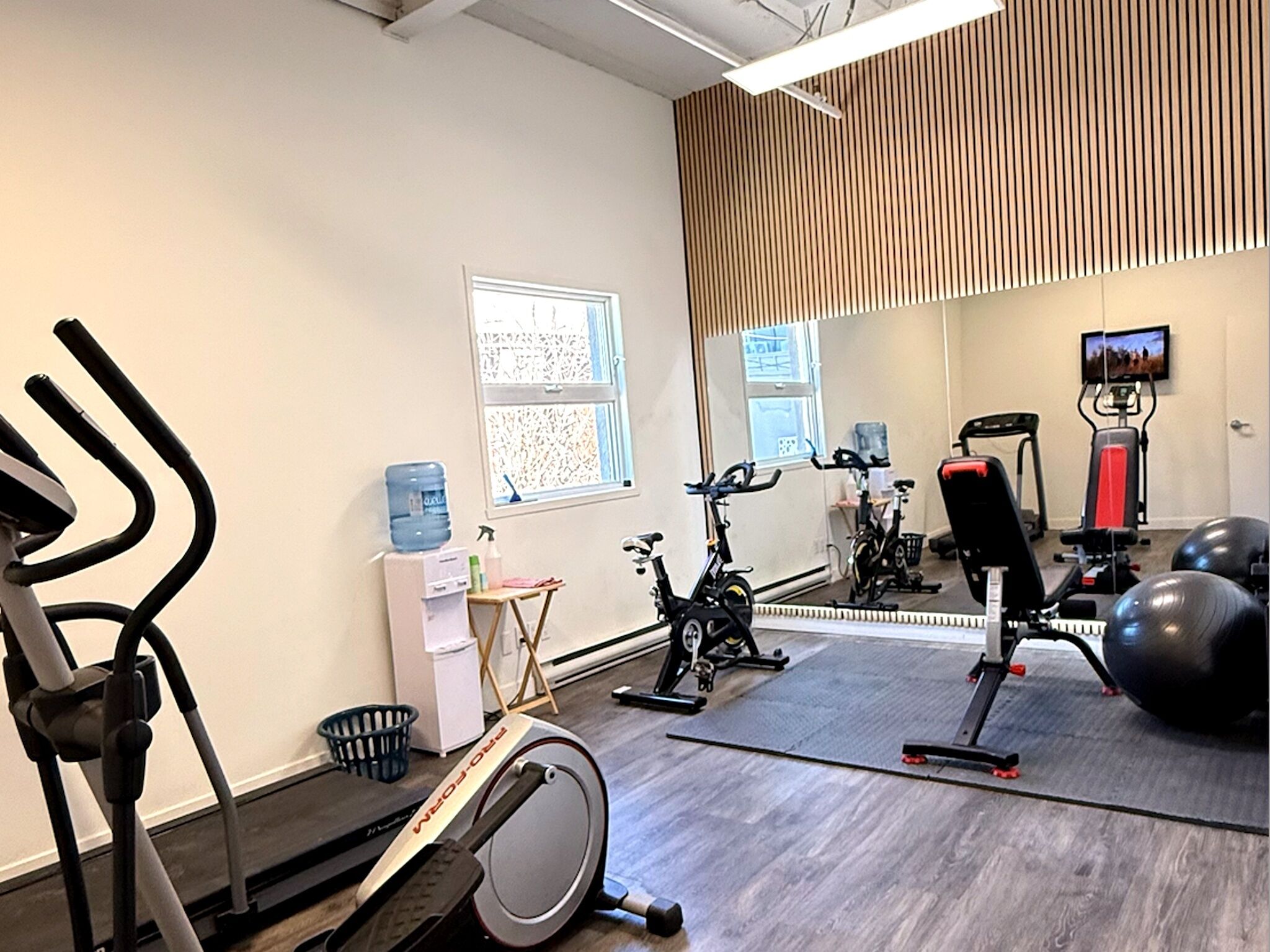 Un gimnasio con equipo de ejercicio, un dispensador de agua, una pequeña mesa con productos de limpieza y un espejo que refleja el equipo.