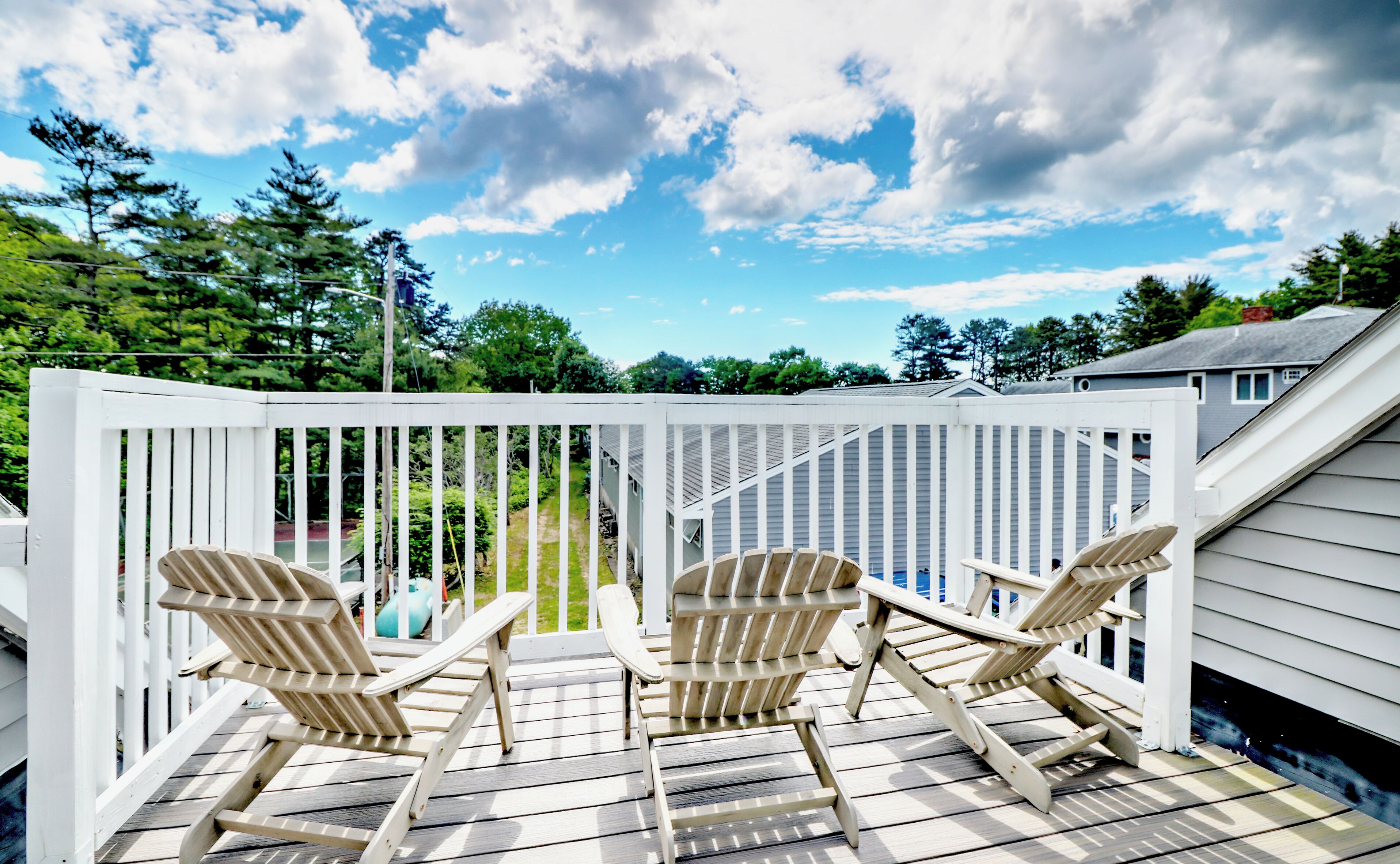 A deck with two Adirondack chairs, a white railing, and a view of a house and trees under a partly cloudy sky.