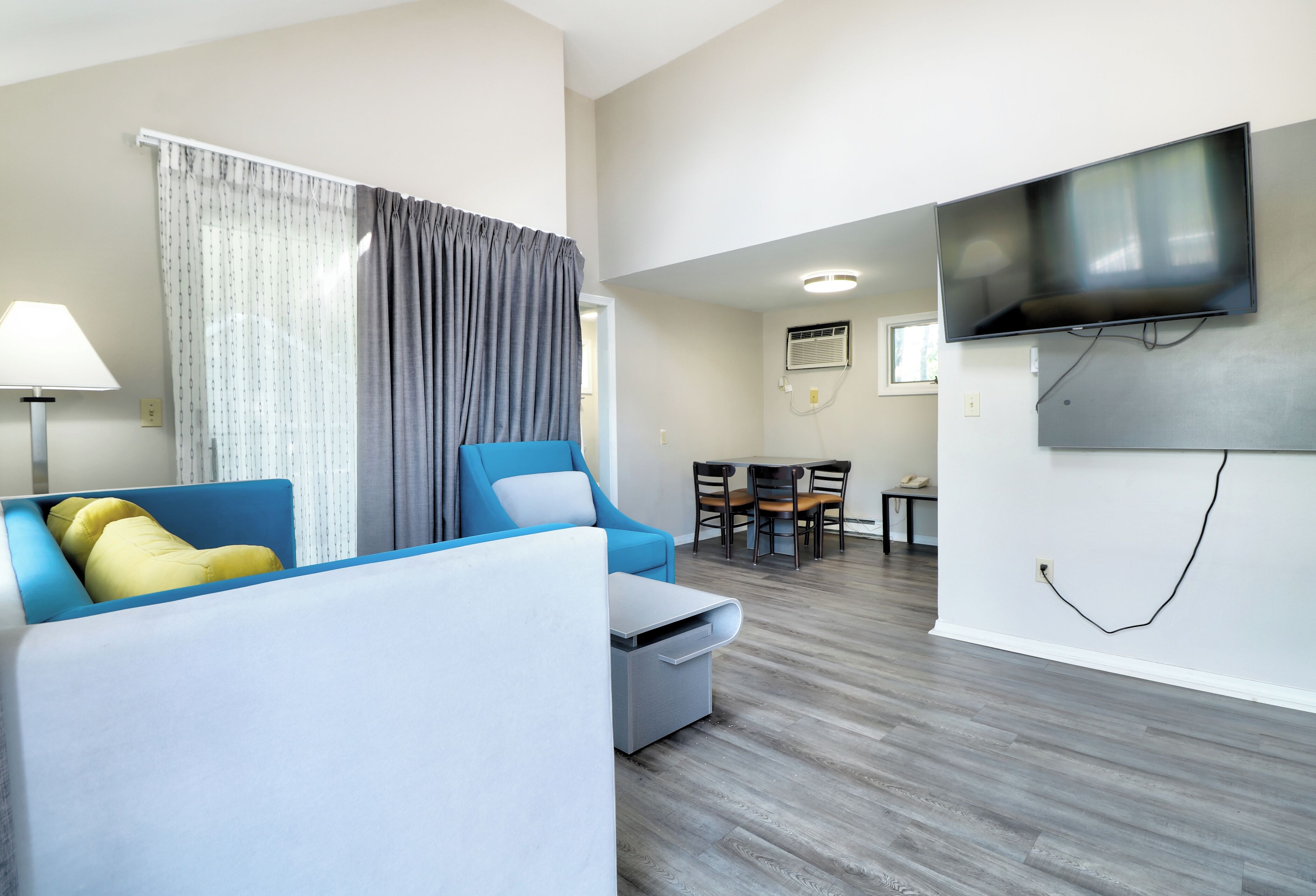 A modern living room with a sofa, a small table, a TV mounted on the wall, and a dining area with chairs and a table.