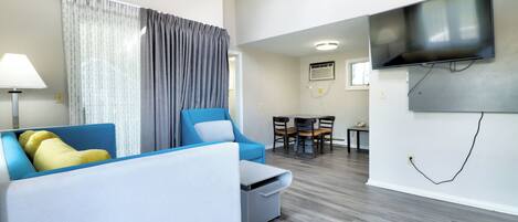 A modern living room with a sofa, a small table, a TV mounted on the wall, and a dining area with chairs and a table.