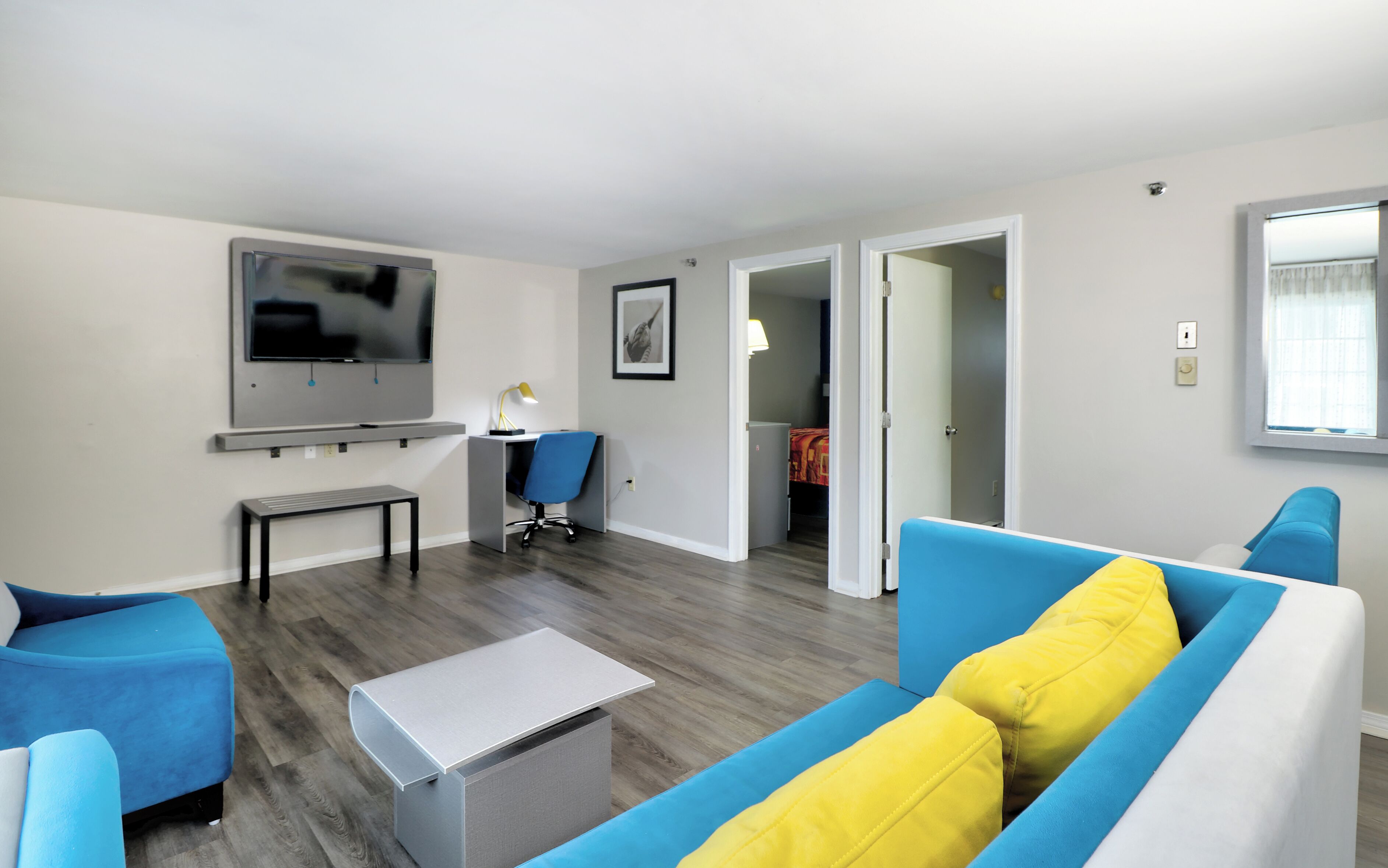 Een moderne woonkamer met een flatscreen televisie, een bureau, een blauwe stoel, een bank met gele kussens en een kleine tafel.