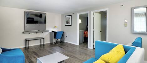Een moderne woonkamer met een flatscreen televisie, een bureau, een blauwe stoel, een bank met gele kussens en een kleine tafel.