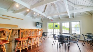Un restaurant avec des chaises et des tables en bois, un téléviseur fixé au mur et de grandes fenêtres ornées d’une enseigne indiquant « OUVERT ».