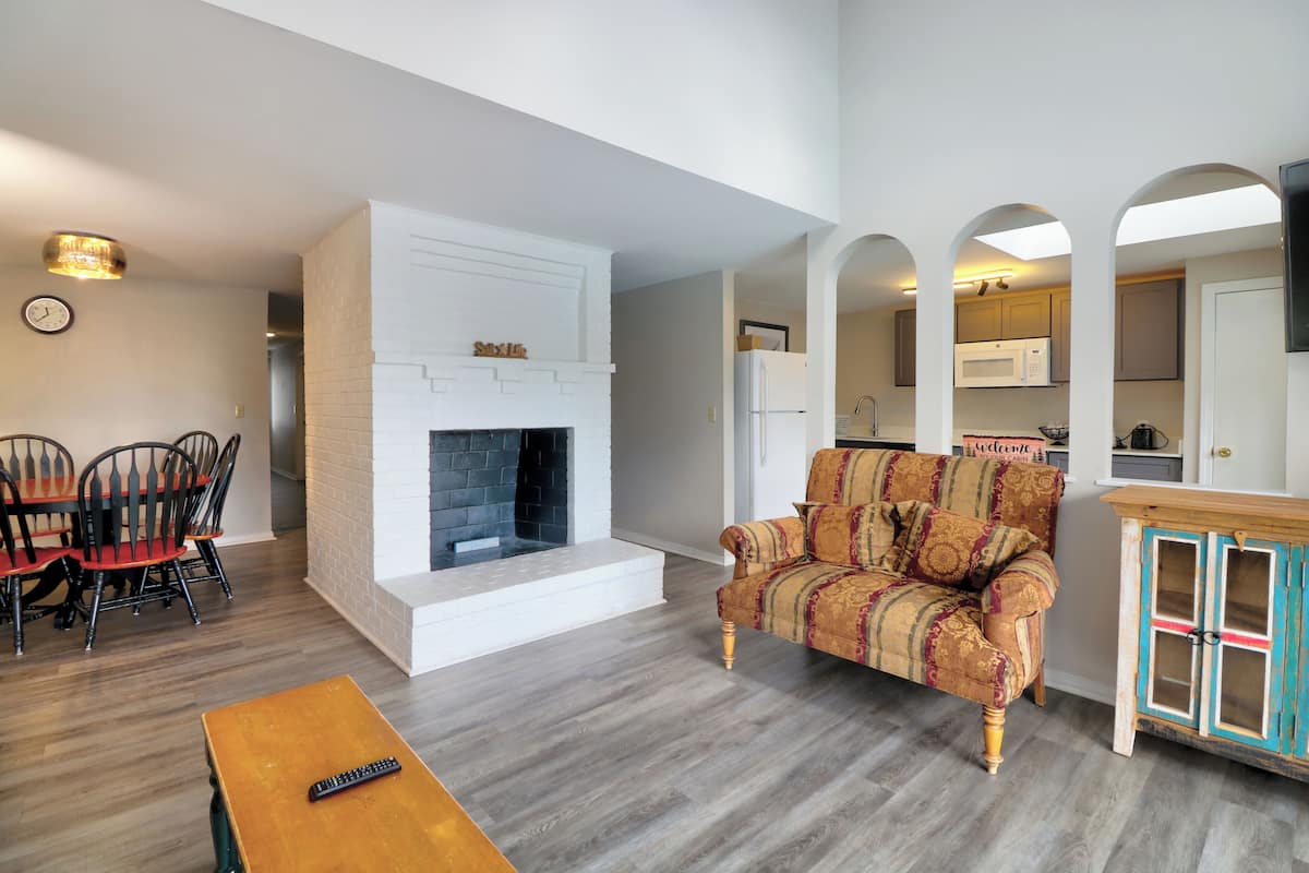 A living room with a fireplace, a sofa, a dining table with chairs, and a kitchen area visible through an archway.