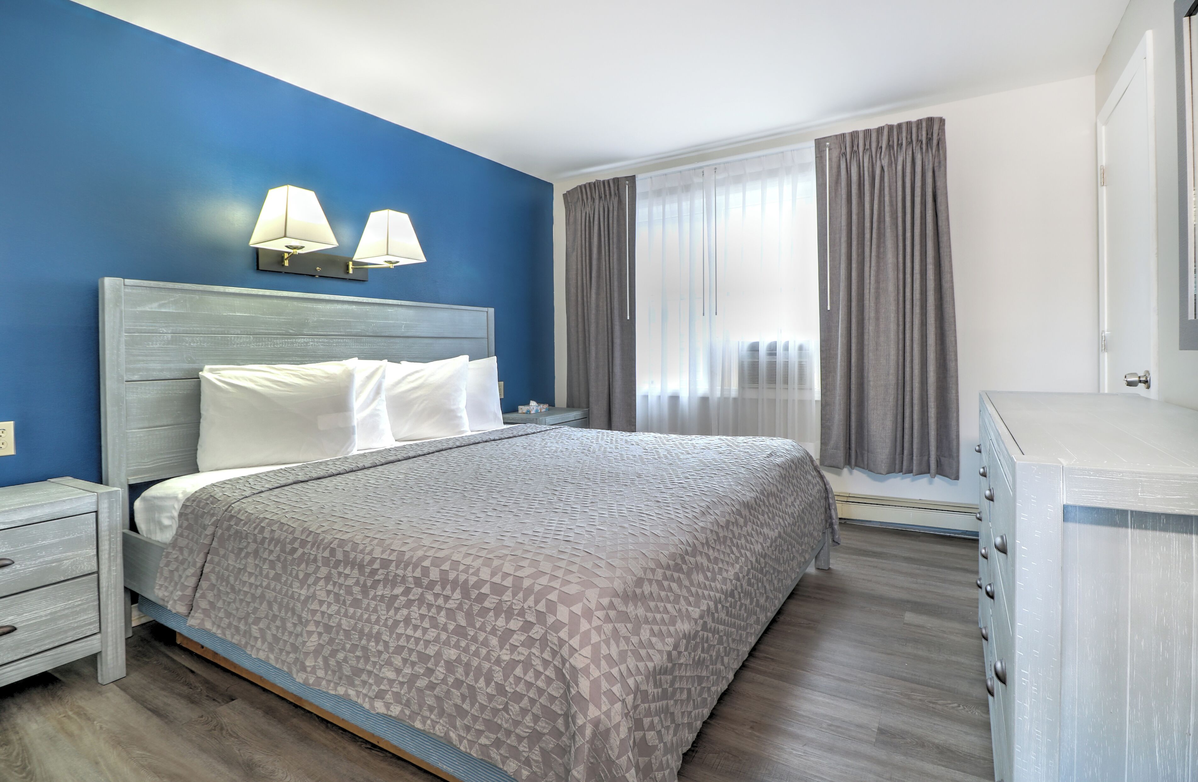 A hotel room with a bed, bedside tables, a dresser, and a window with curtains.