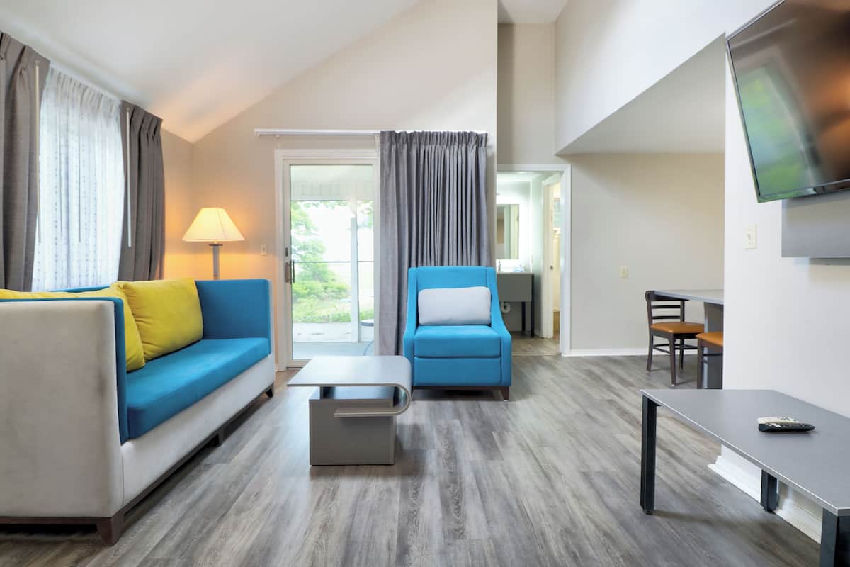 A modern hotel room with a sofa, armchair, coffee table, TV, and a dining area.