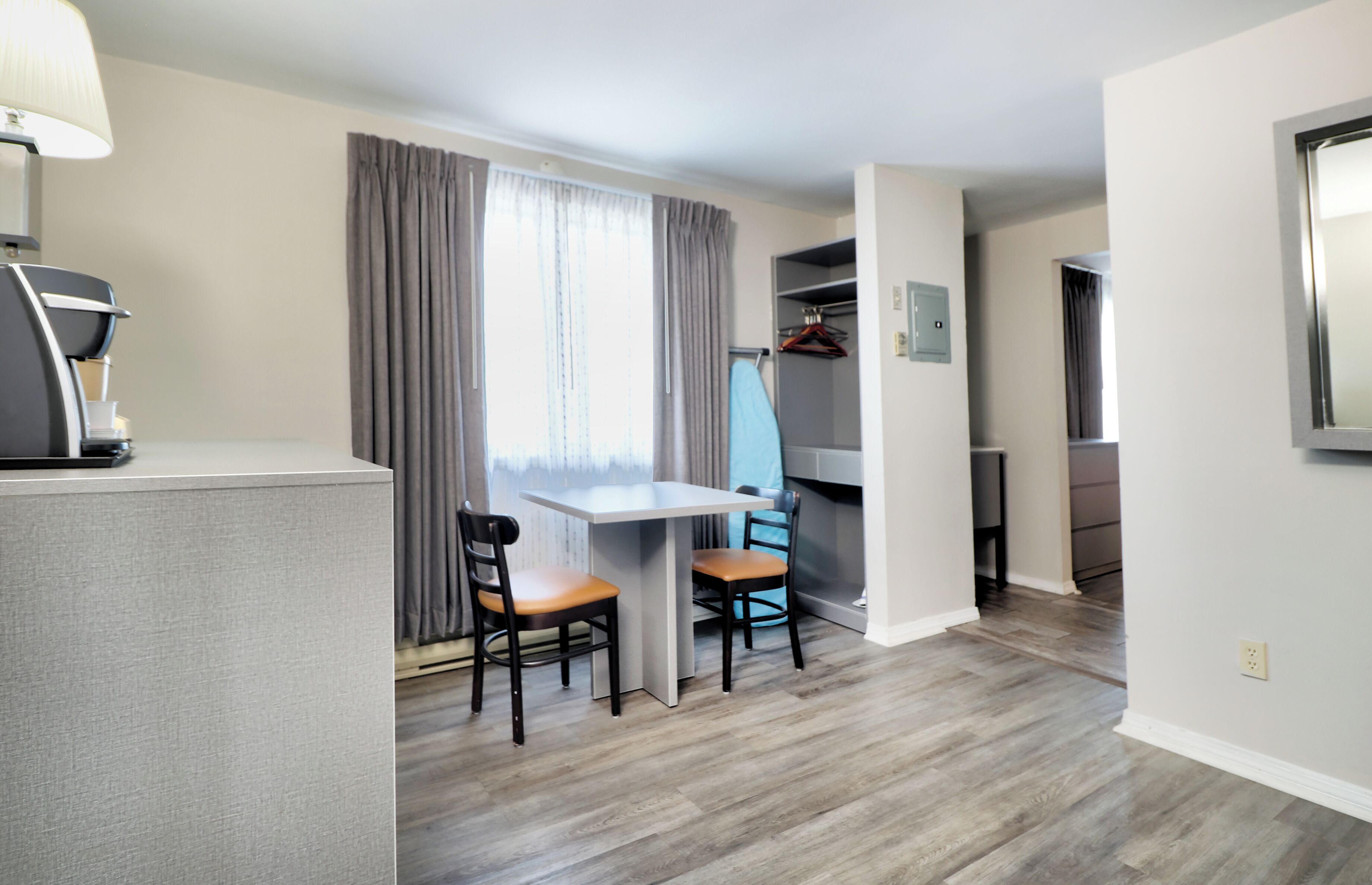 A modern hotel room with a kitchenette, a dining area with a table and chairs, a wardrobe, and a large window with curtains.