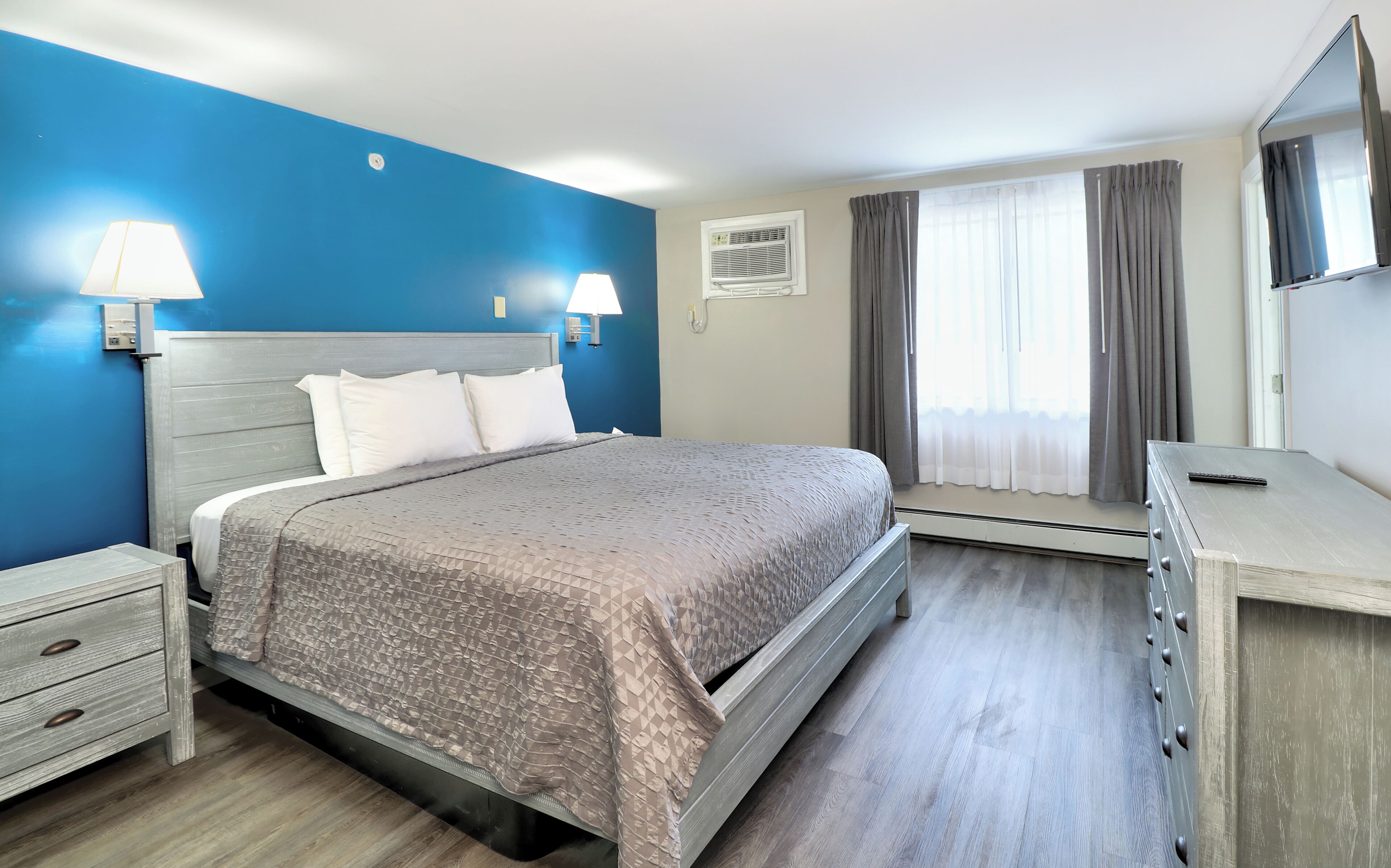 Ein Hotelzimmer mit Bett, Nachttischen, einem Schminktisch, einem Fenster mit Vorhängen und einer Klimaanlage.