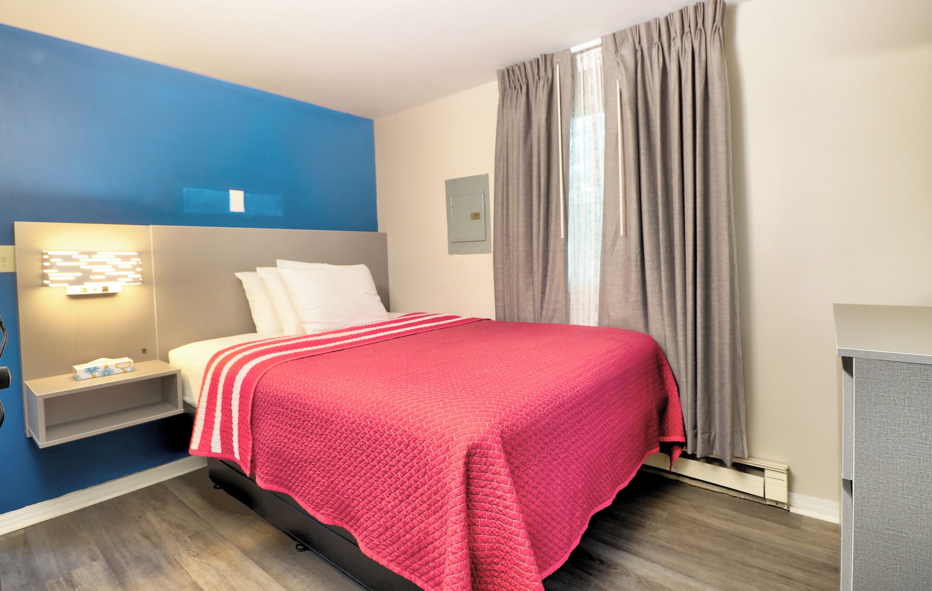 Ein Hotelzimmer mit Bett, Nachttisch, Wandleuchte, einem Fenster mit Vorhängen und einem eingebauten Schrank.