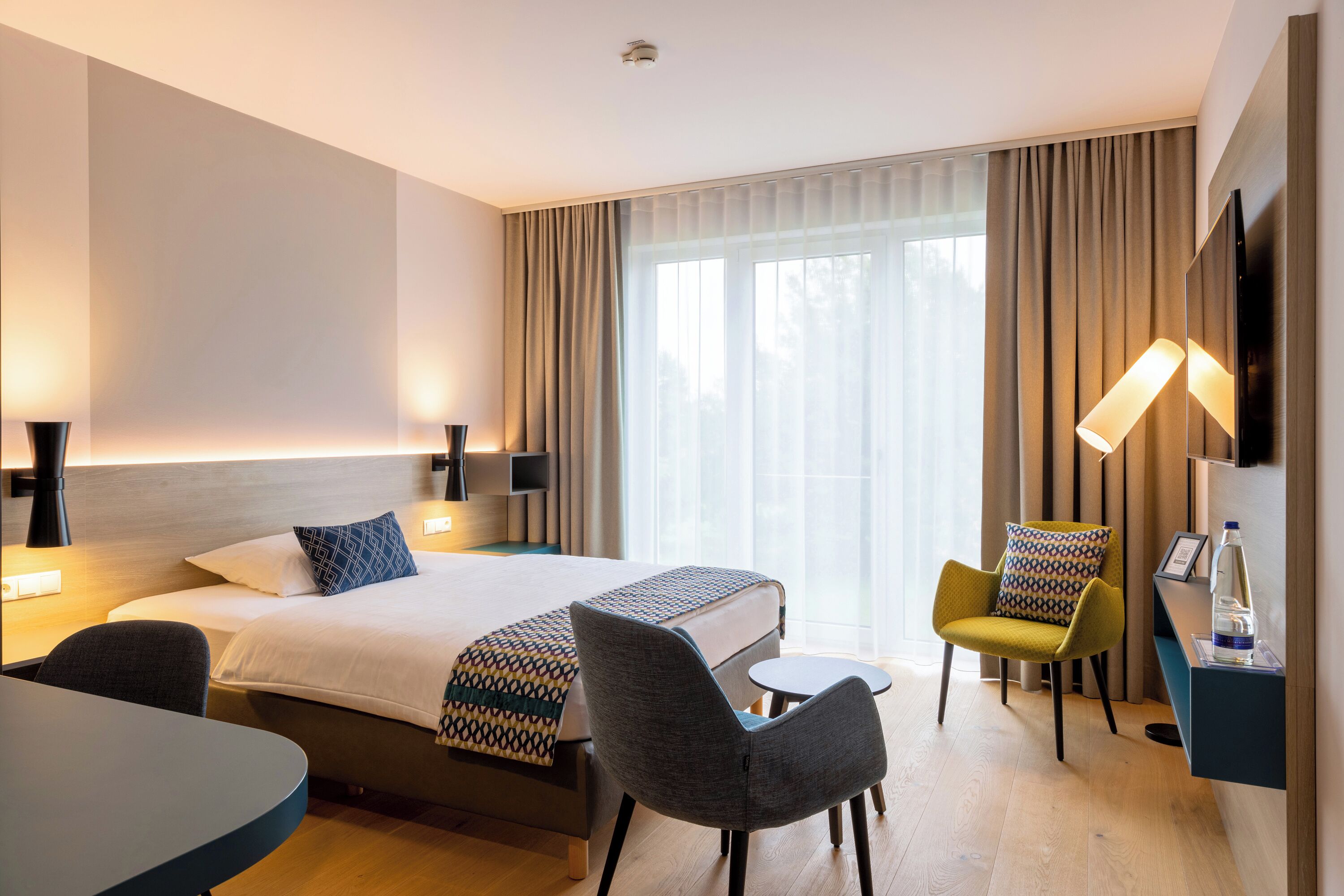 Ein Hotelzimmer mit einem großen Bett, zwei Nachttischen, einem Schreibtisch, zwei Stühlen, einem Fernseher und einem großen Fenster mit Vorhängen.